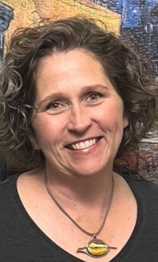 Smiling person with shoulder-length curly brown hair wearing a dark shirt and a pendant necklace, against a textured wall.