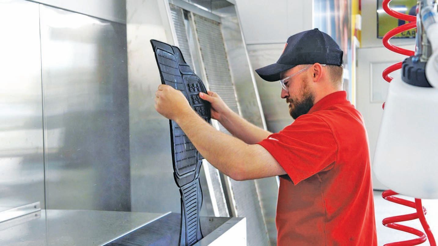 A worker in a red shirt cleans a dark rubber floor mat at a professional car wash station.