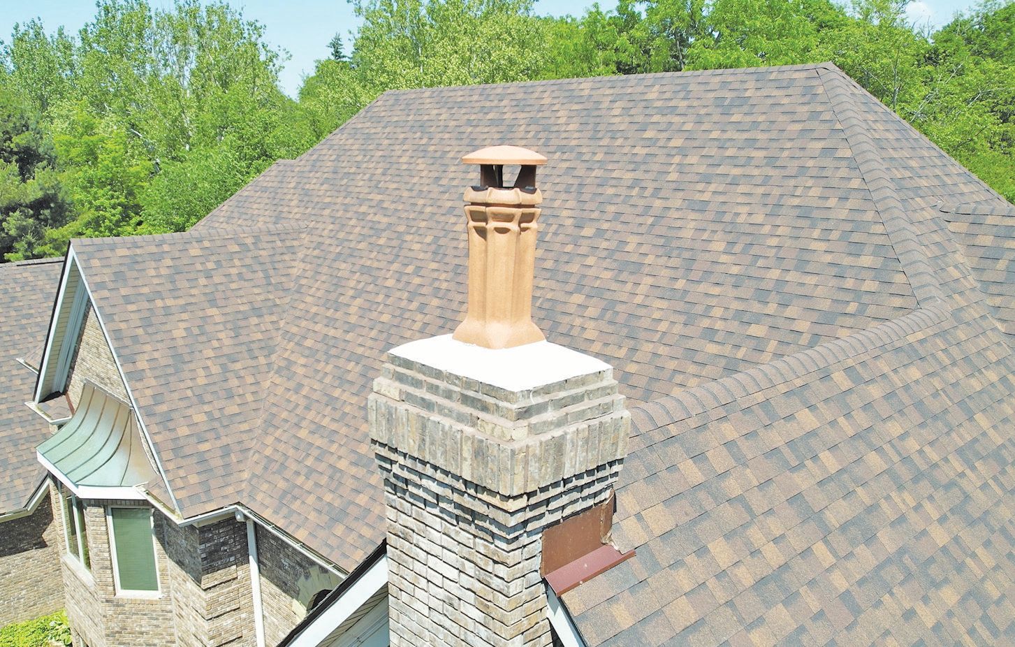 A brick chimney with a terracotta chimney pot stands on a residential roof covered in dark, multi-toned asphalt shingles.