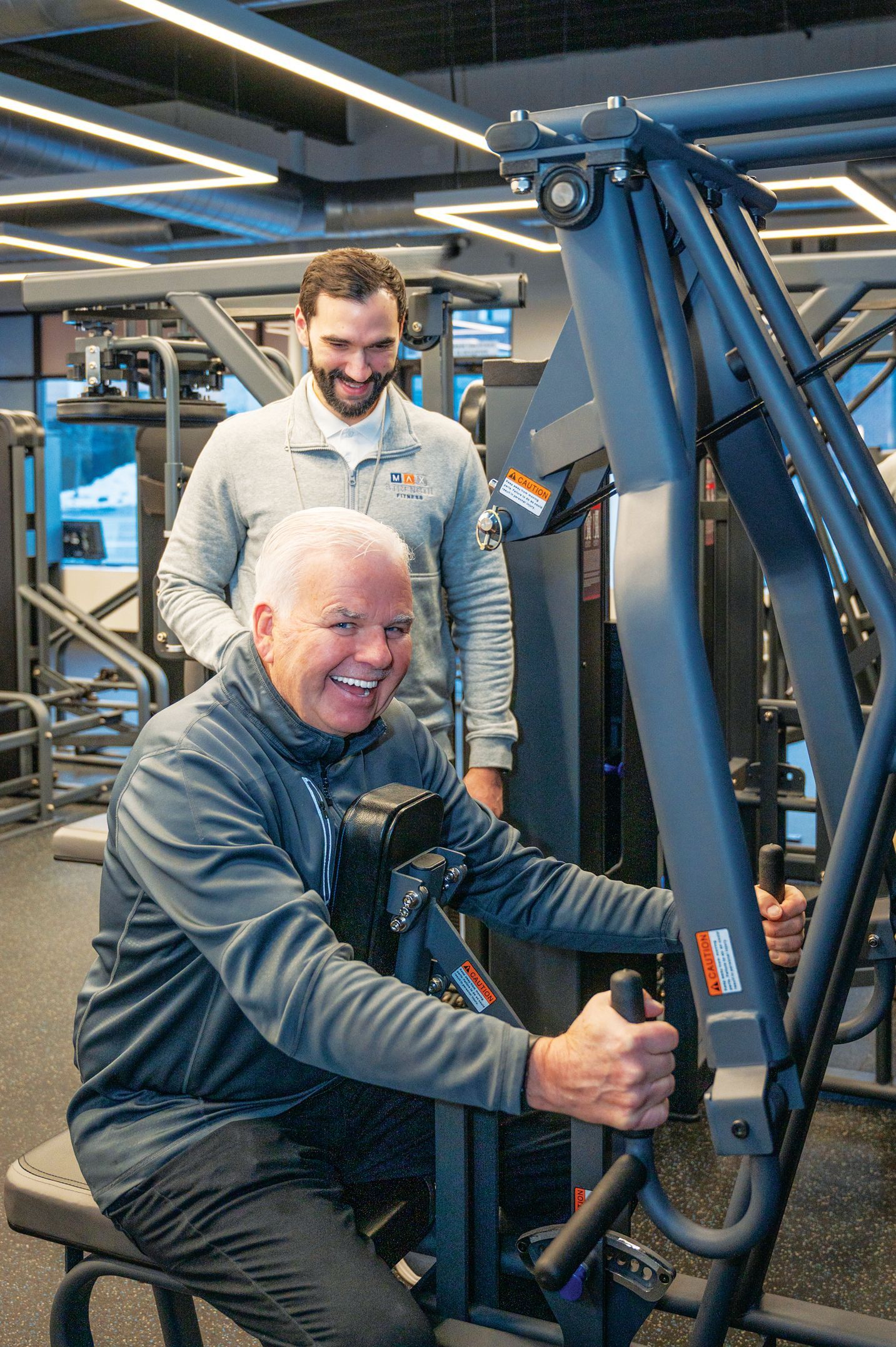 A gym instructor smiles while supervising a person exercising on a chest press machine in a modern fitness center.