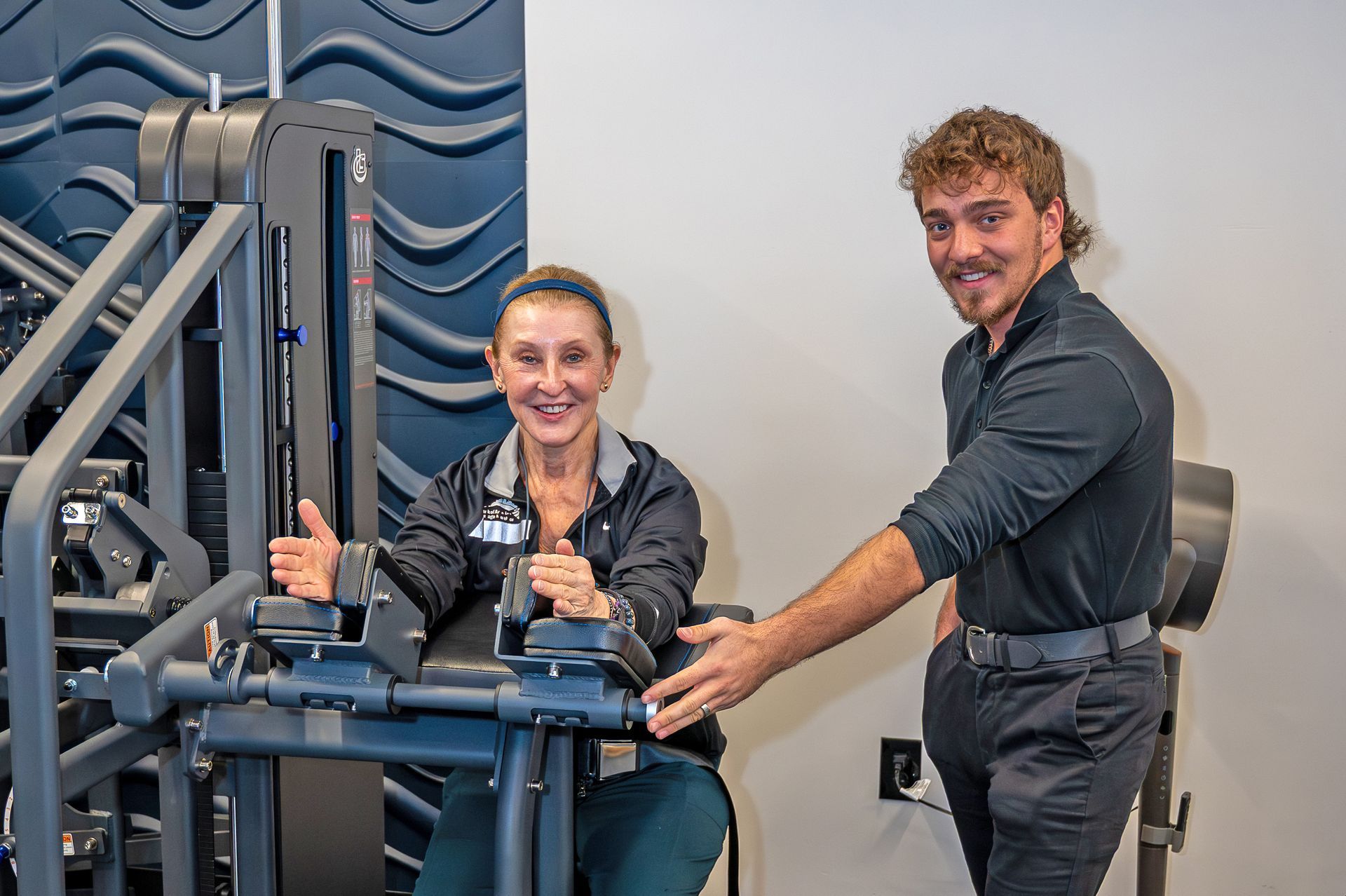 A smiling person uses a gym machine while a trainer gestures toward the equipment in a modern exercise room.