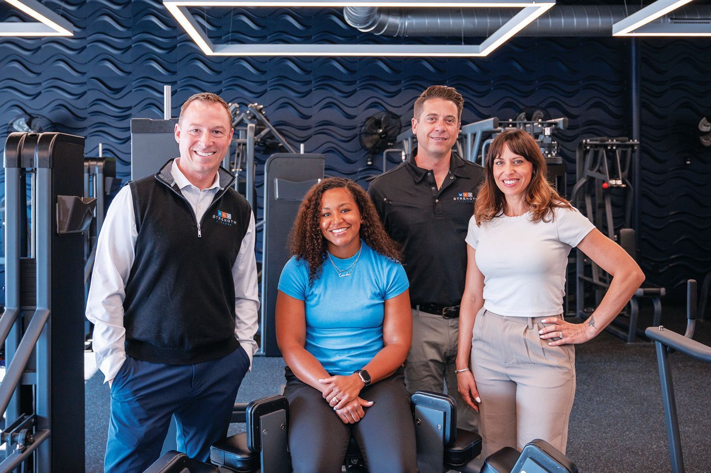 Four people stand and sit in a modern gym with dark, textured walls, smiling toward the camera.