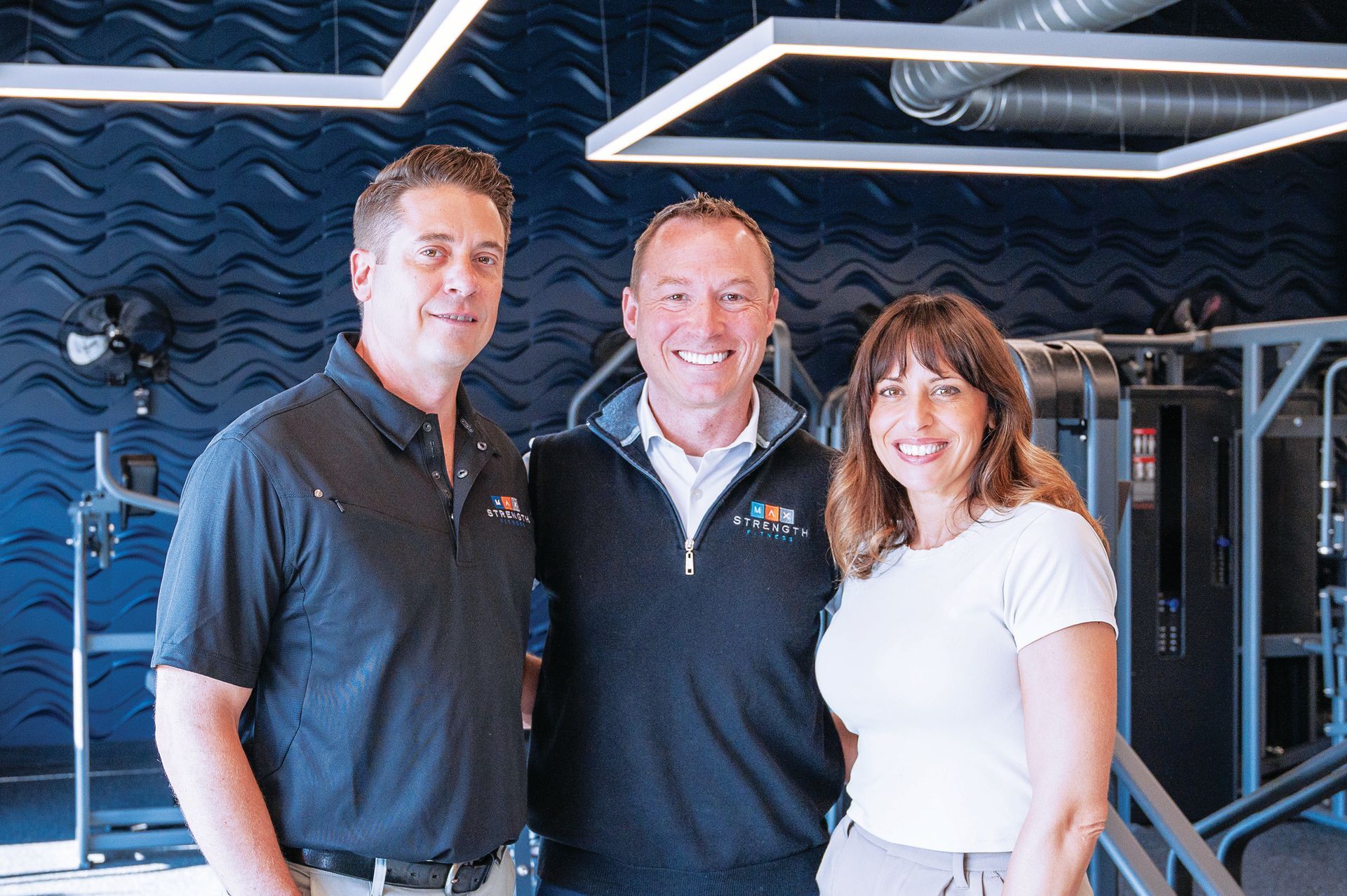Three people smiling side-by-side in a modern gym with blue textured walls and fitness equipment in the background.
