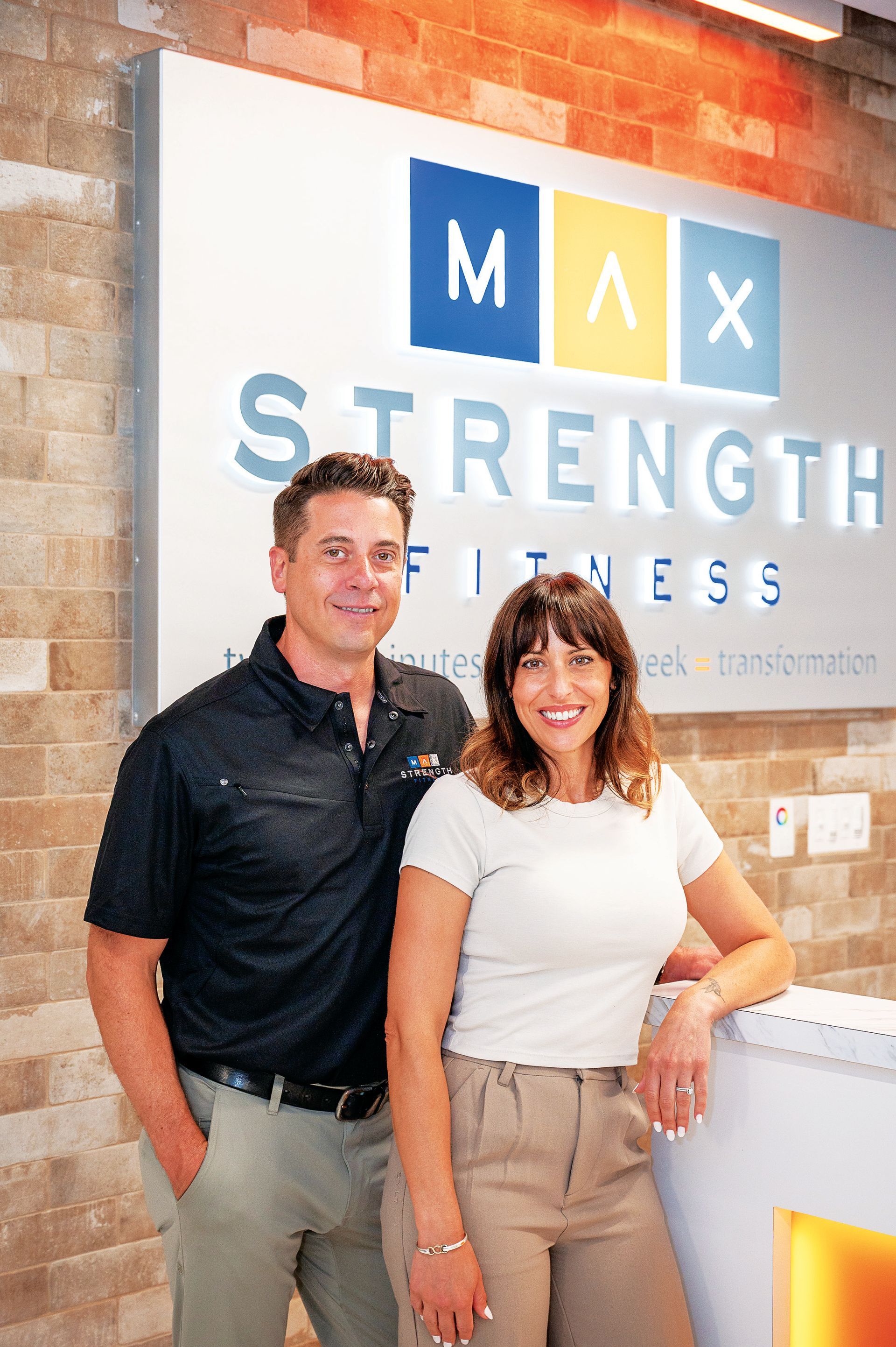 A man and a woman pose smiling in front of a Max Strength Fitness sign against a brick wall.