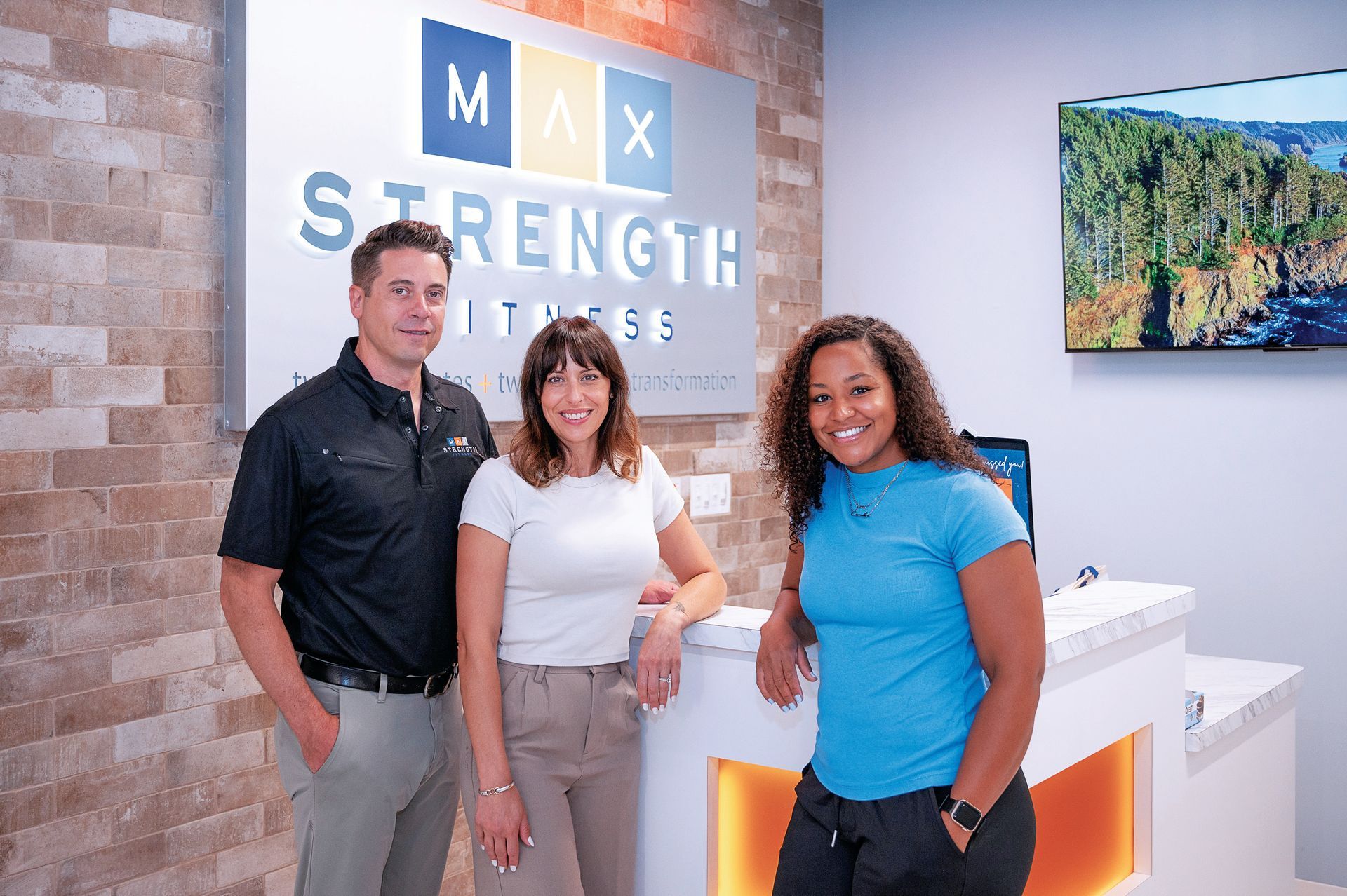 Three people standing behind a reception desk in a gym titled Max Strength Fitness with a stone wall and digital screen.