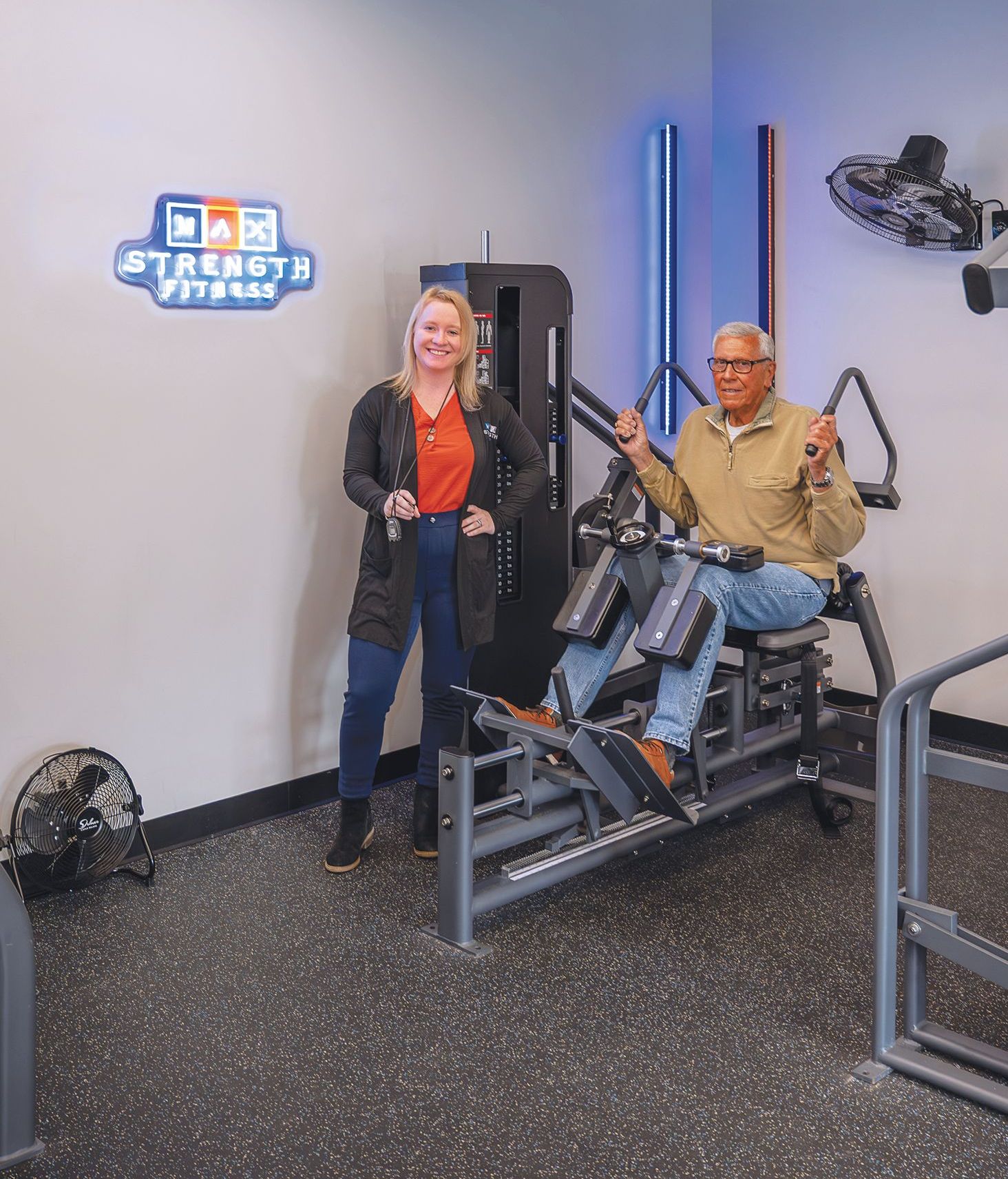 A person standing next to an individual seated on a weight machine in a gym with a Strength Fit gym logo on the wall.