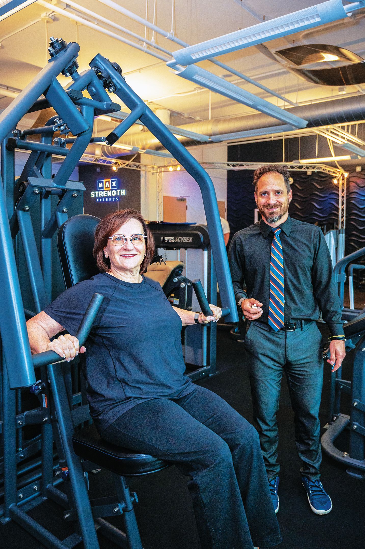 A person sits at a gym chest press machine while another person stands nearby, both smiling in a gym setting.