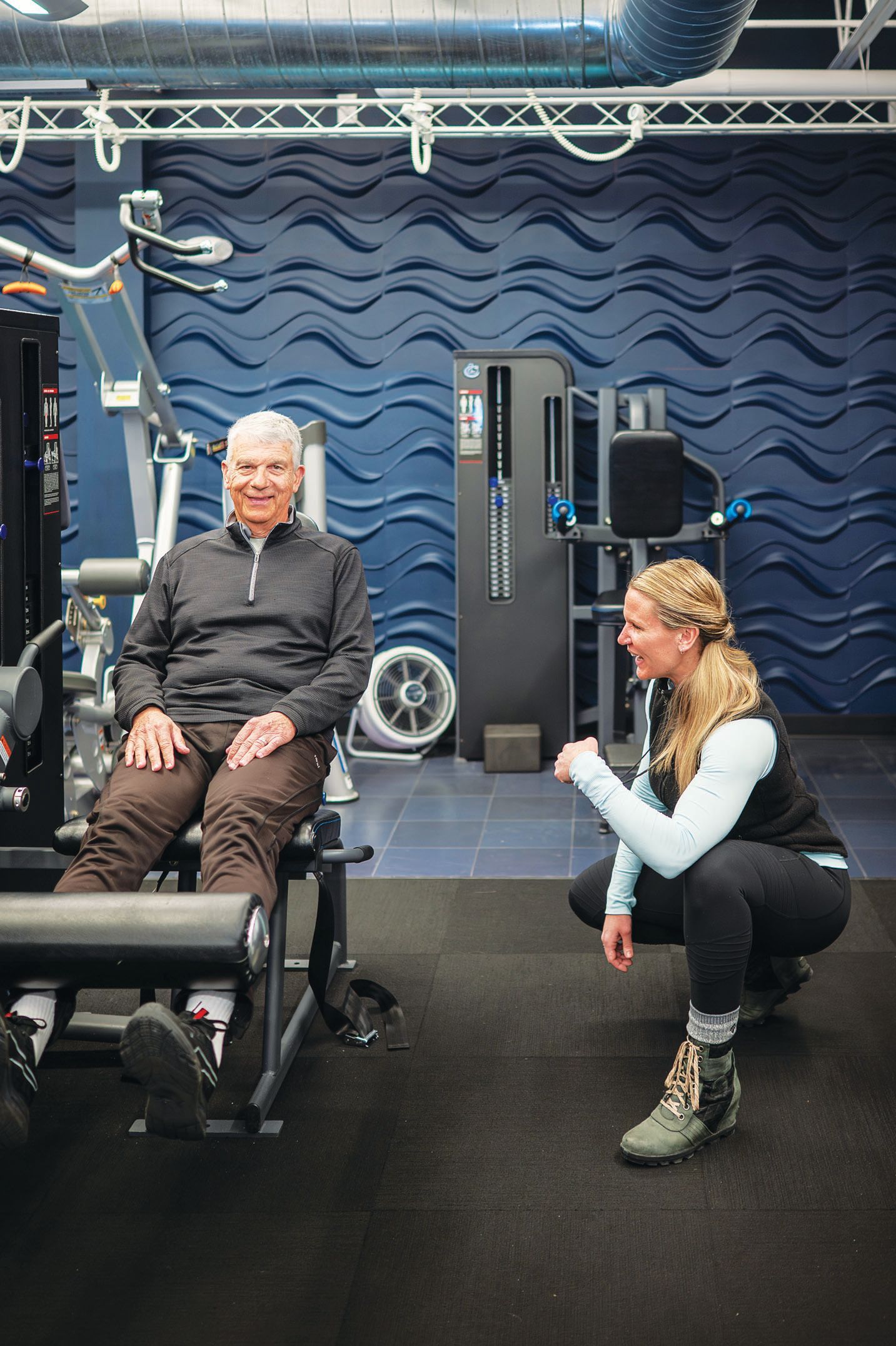 A smiling man sits at a gym machine while a fitness professional crouches nearby, coaching him during a workout.