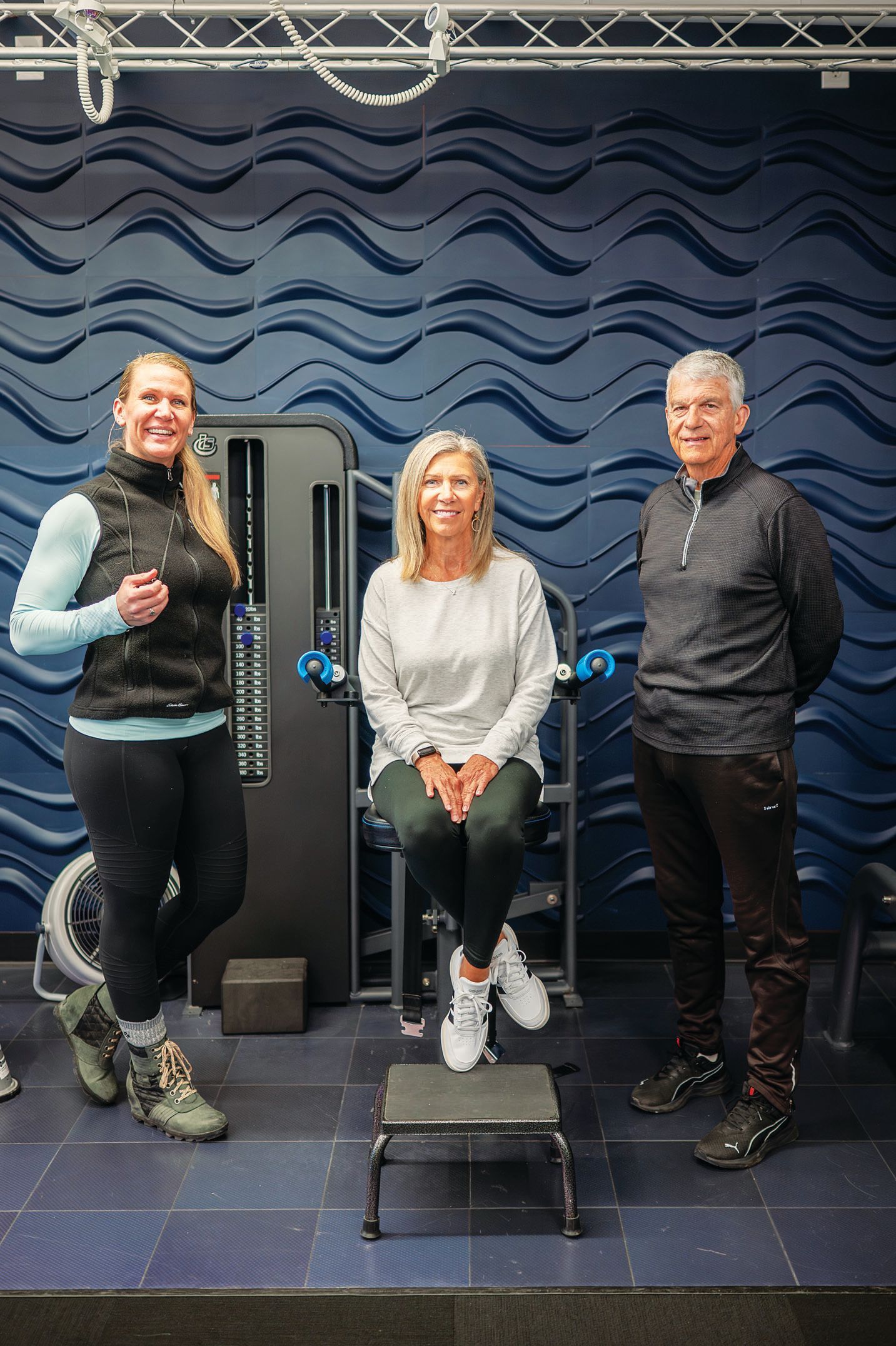 Three people stand and sit in a gym setting with blue textured walls, posing near exercise equipment.