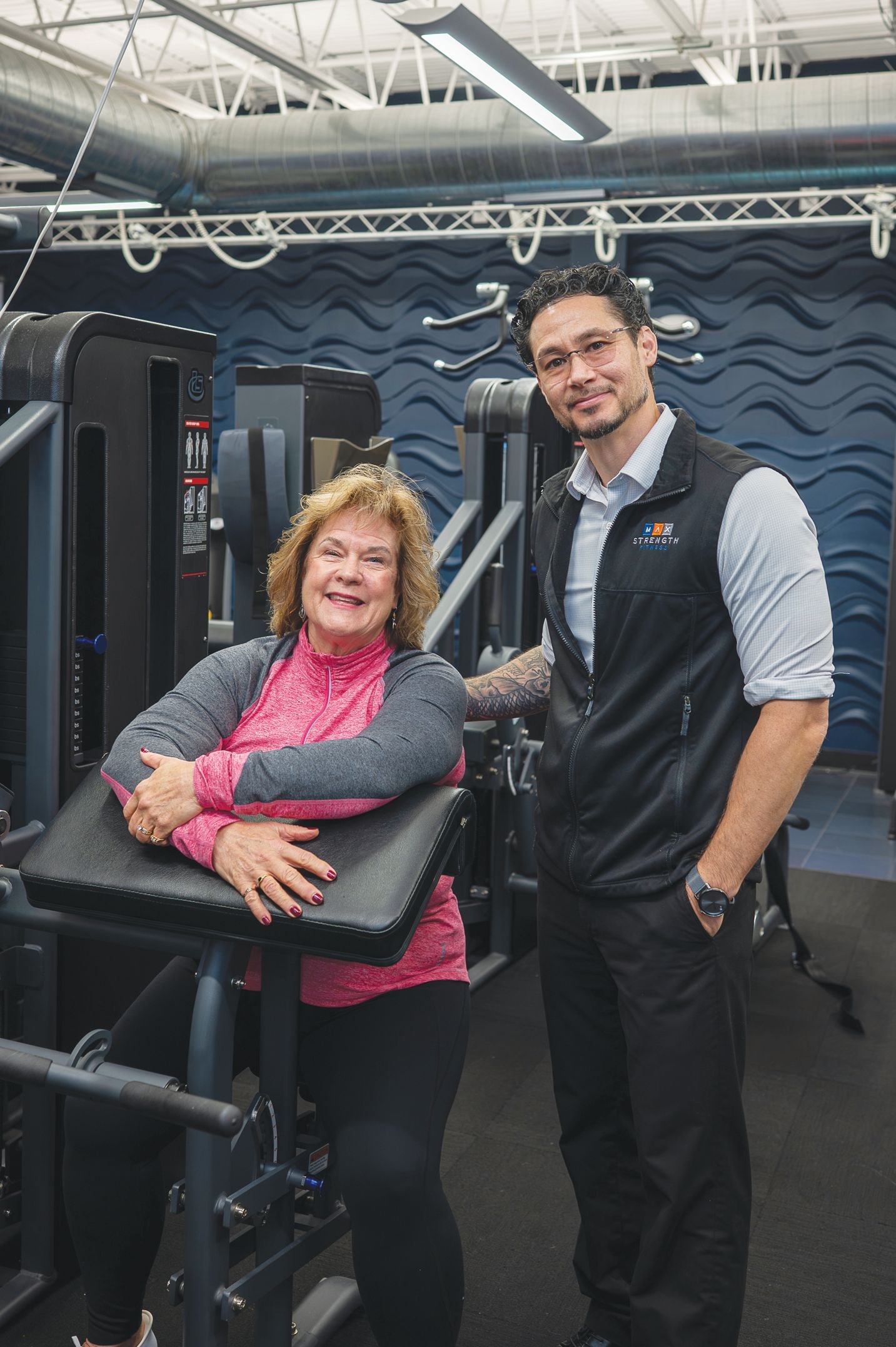 A smiling woman sits at a gym machine with a trainer standing next to her in a fitness center.