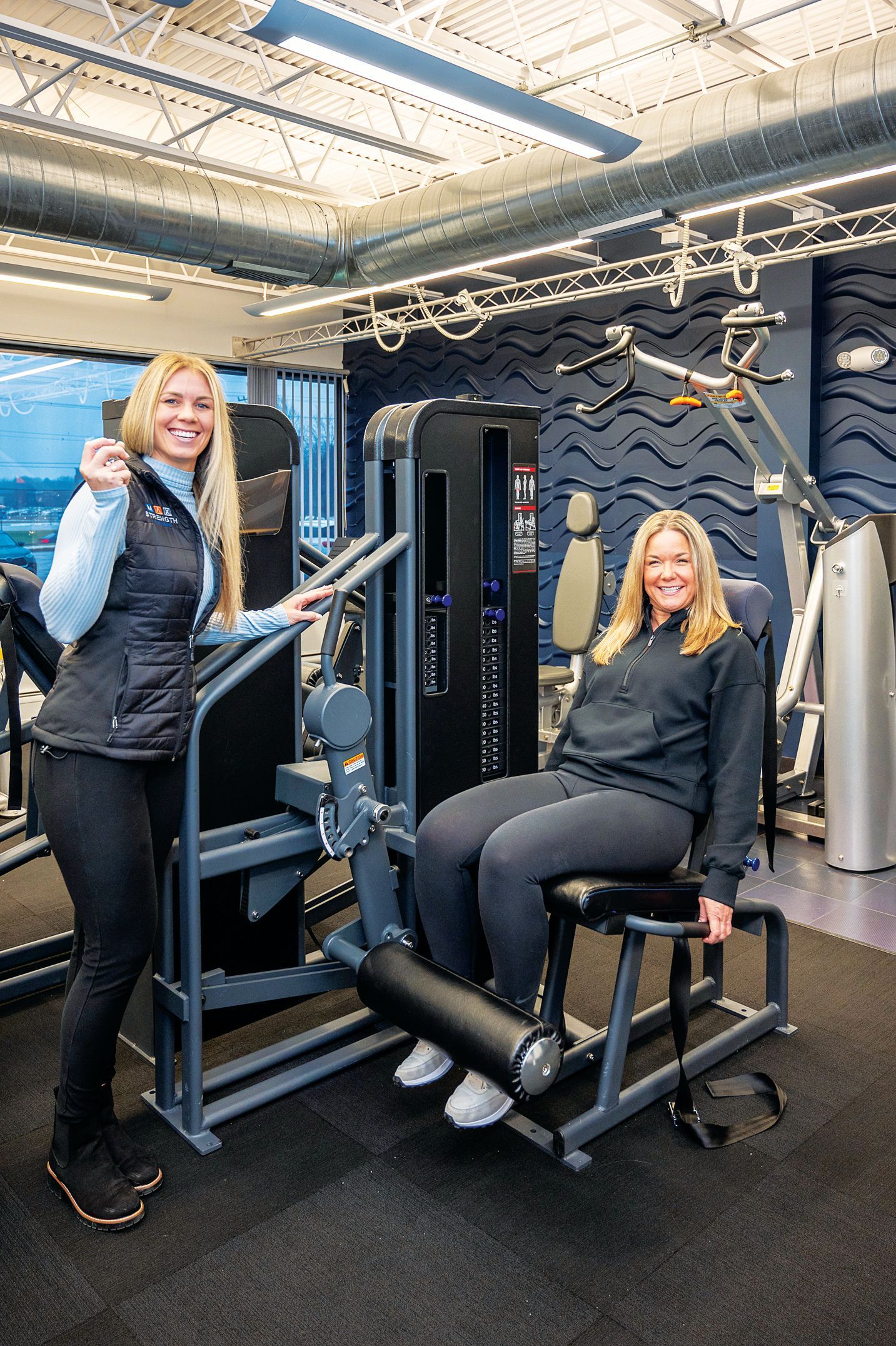 Two smiling people in a modern gym setting, one standing next to and one seated on a leg extension weight machine.