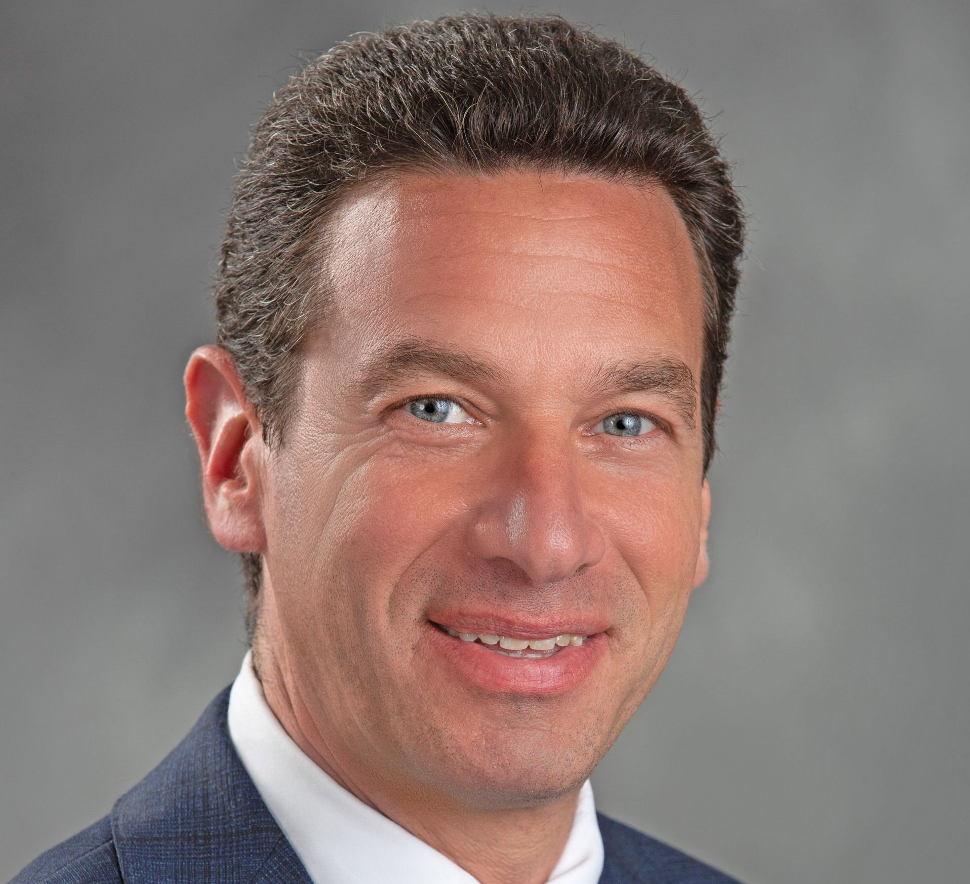 Headshot of a man with short brown hair wearing a blue suit jacket and white collared shirt against a gray background.