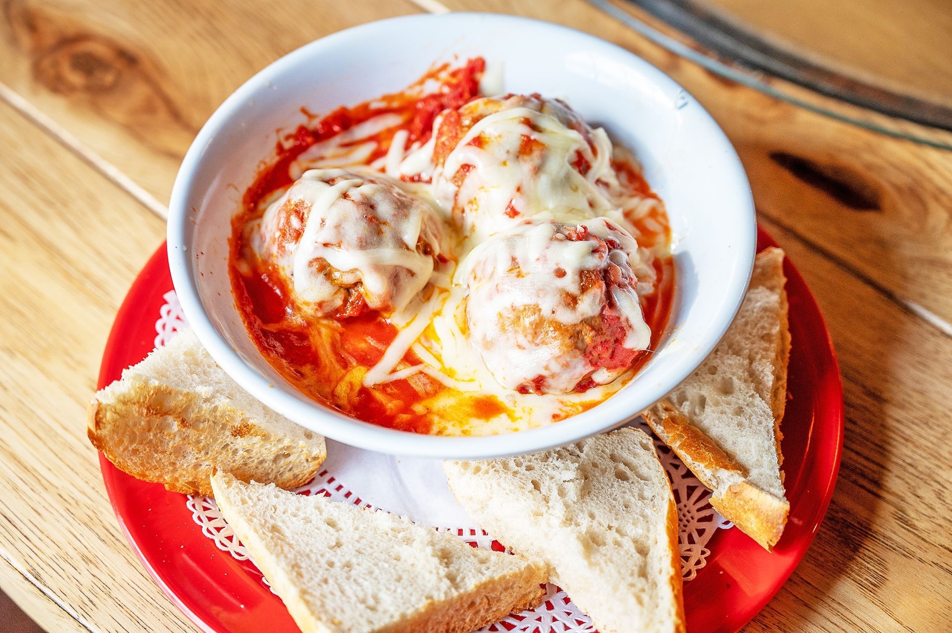 A white bowl of meatballs in red sauce topped with melted cheese, served with crusty bread on a red plate on a wood table.