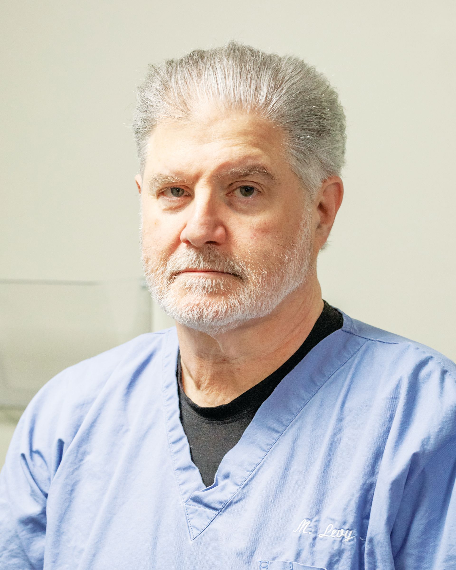 A person with gray hair and a beard, wearing a blue medical scrub top and a black undershirt, looking at the camera.