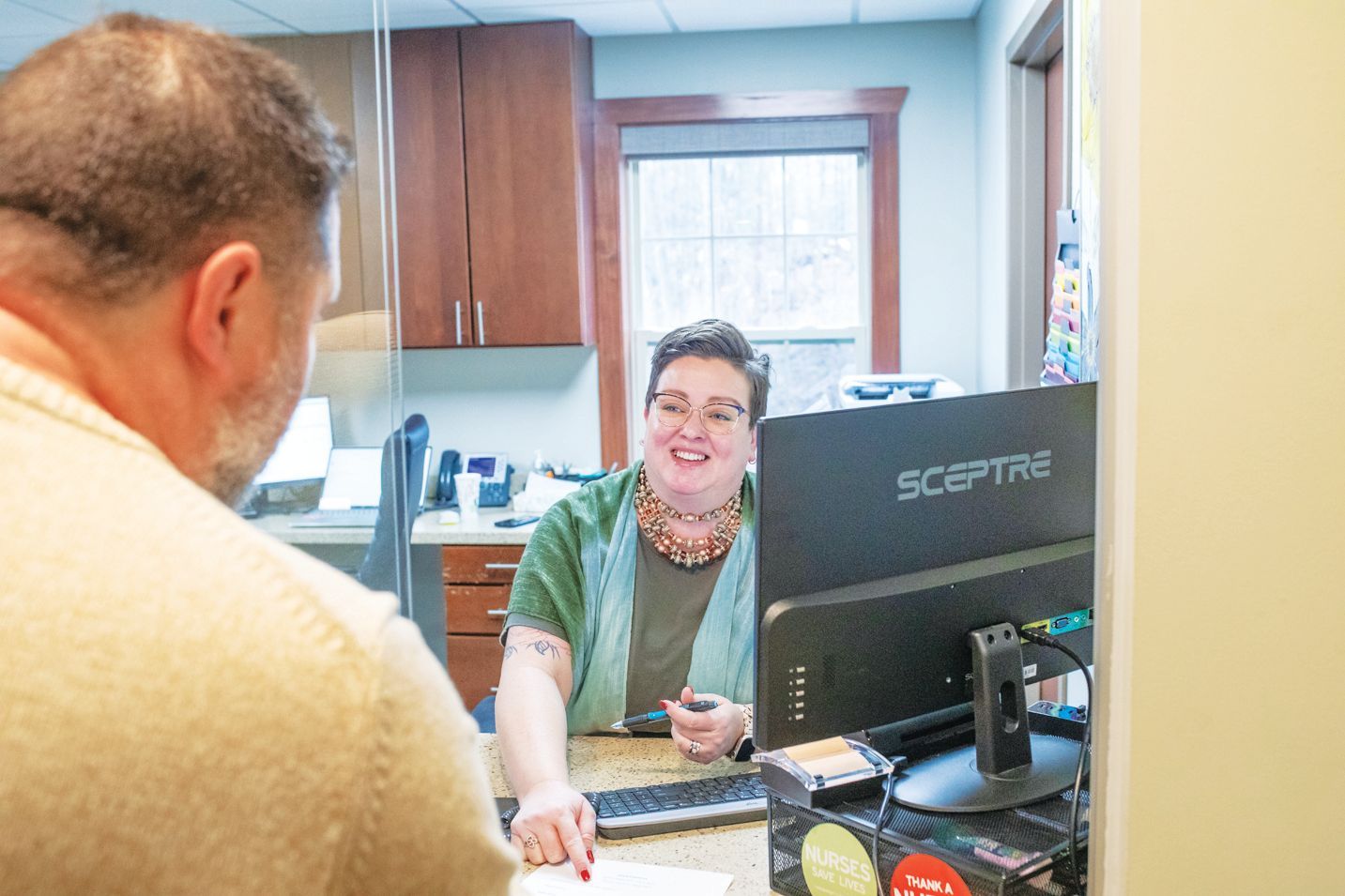 A receptionist in a green shirt smiles and points at a document for a visitor at a service desk.
