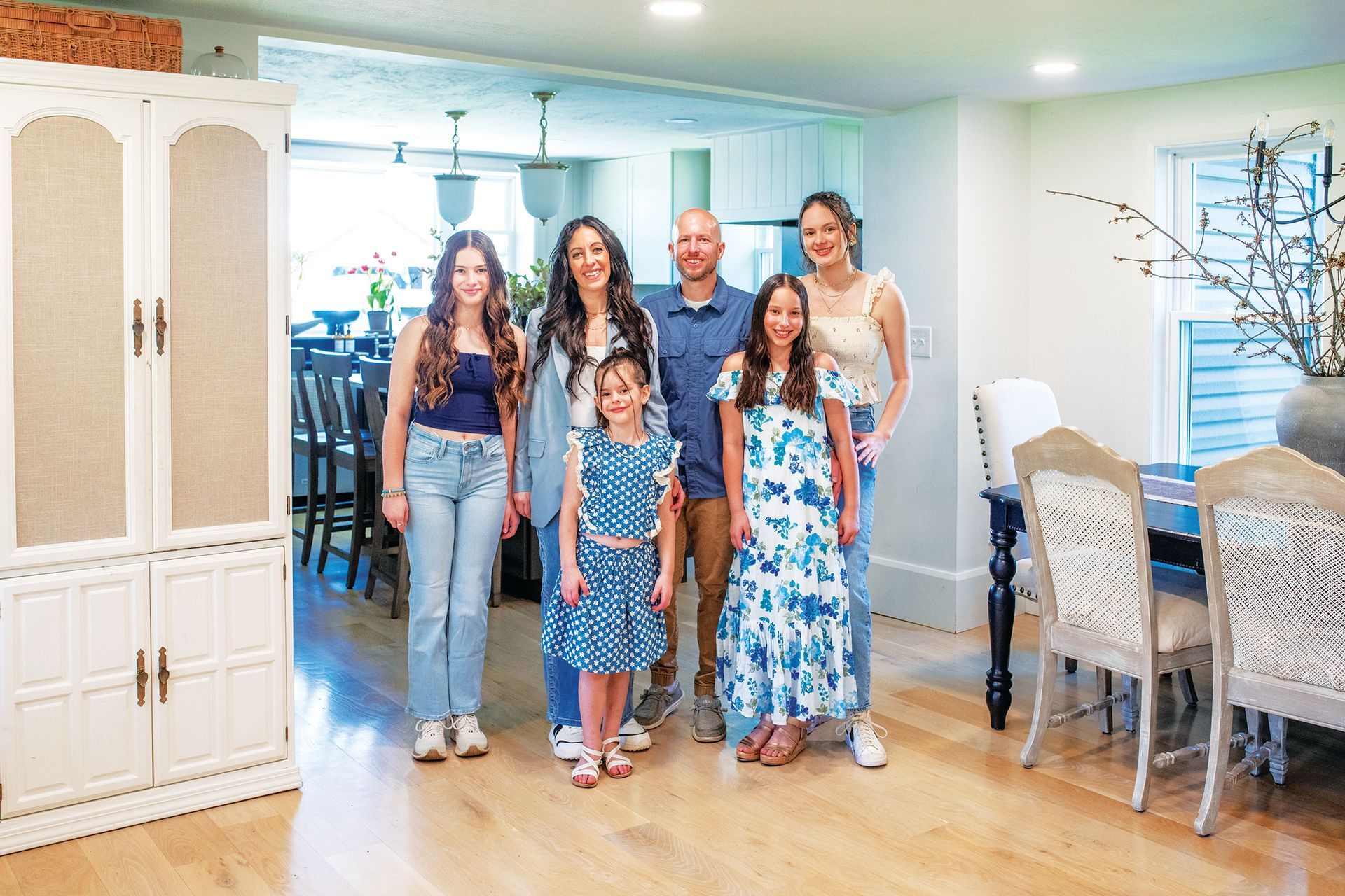 A family of six stands together in a bright, modern dining room with light hardwood floors and a white armoire.