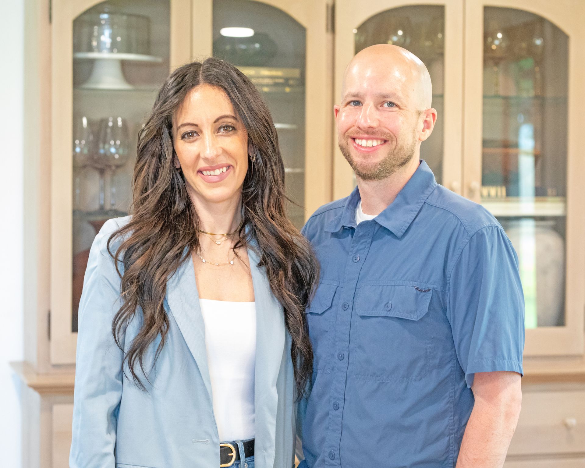 A smiling couple stands side-by-side in front of a light-colored wooden cabinet with glass-paneled doors.