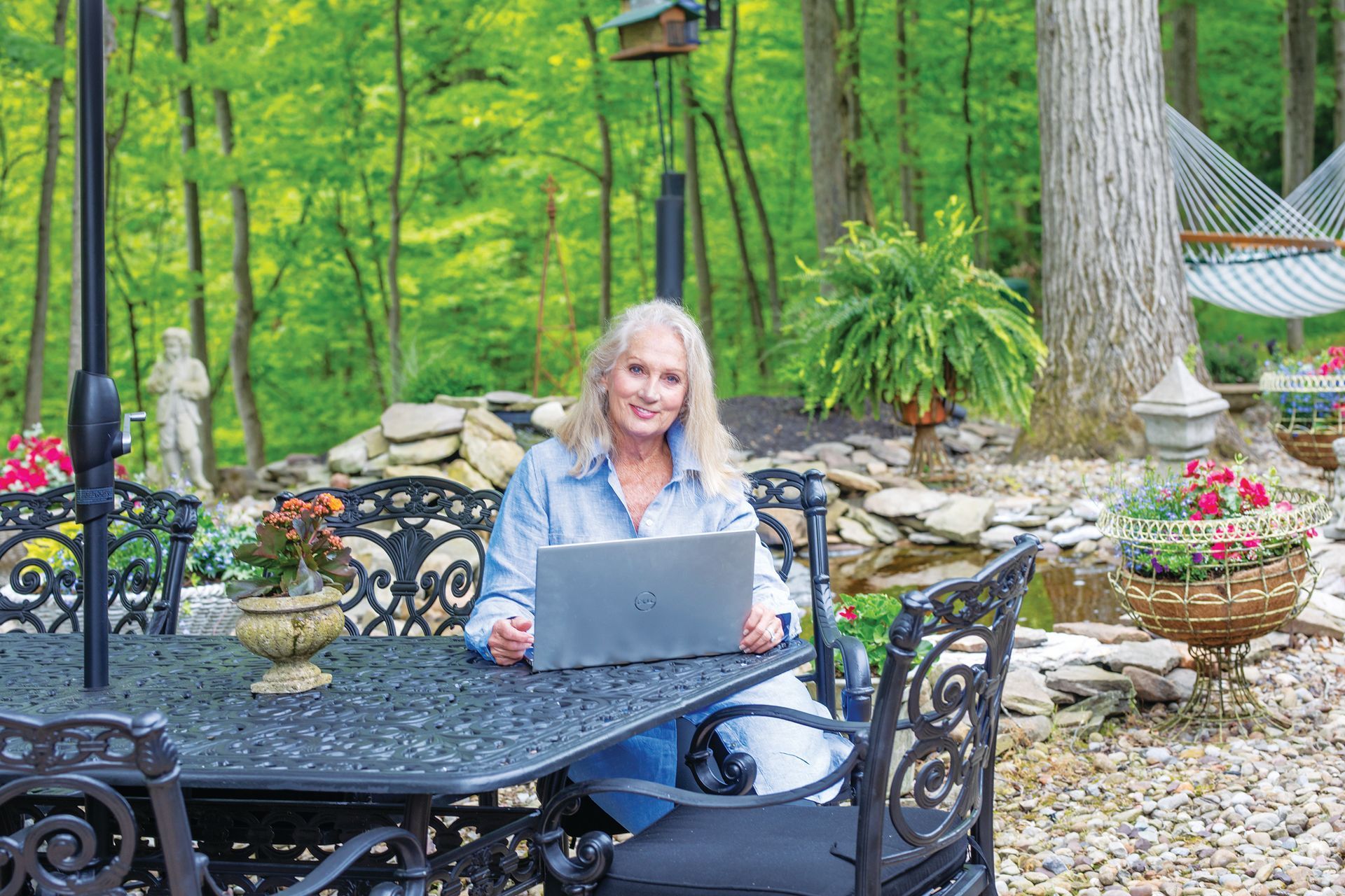 A person sits at an ornate black patio table in a wooded garden, working on a laptop computer.