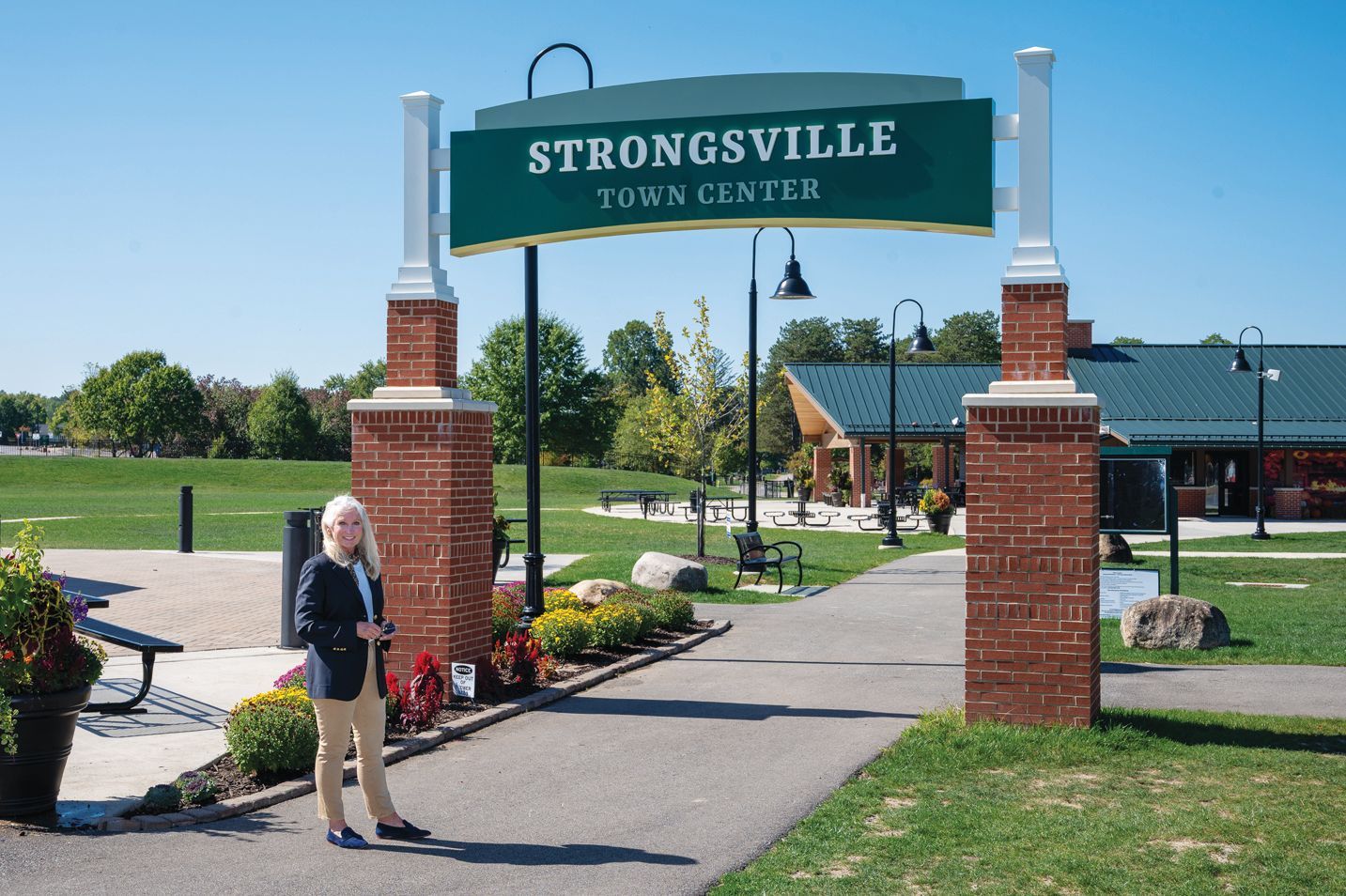 A person stands under the Strongsville Town Center archway on a sunny day with green landscaping and a building in view.