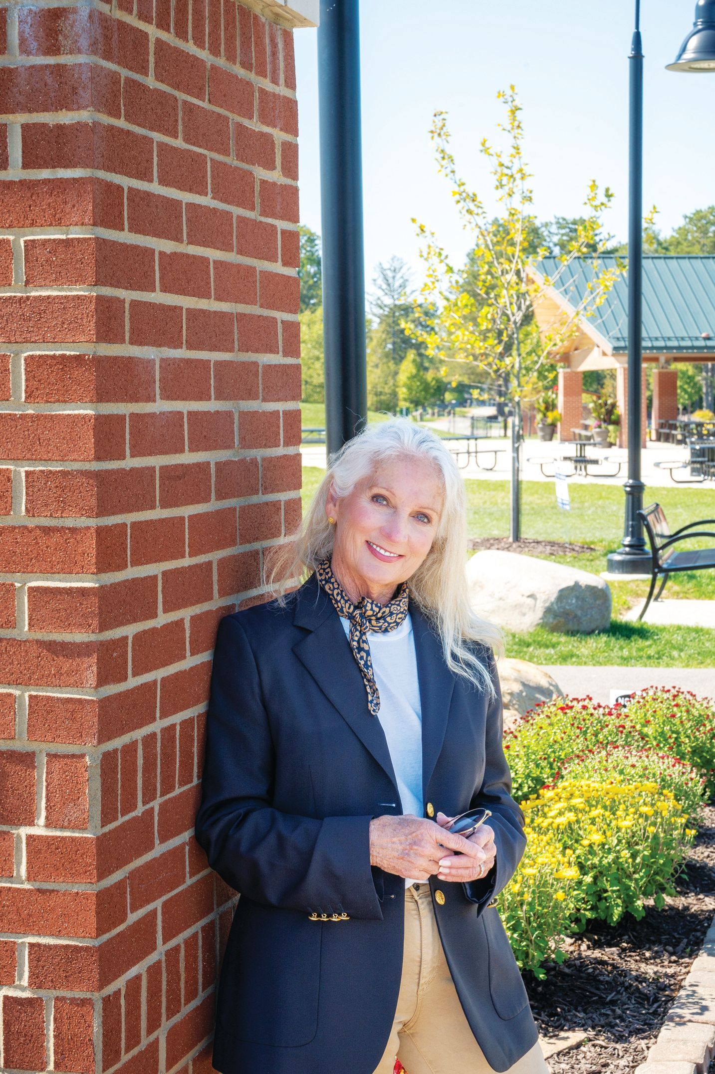 A person leans against a brick pillar outdoors, wearing a navy blazer, light top, and patterned scarf, smiling at the camera.