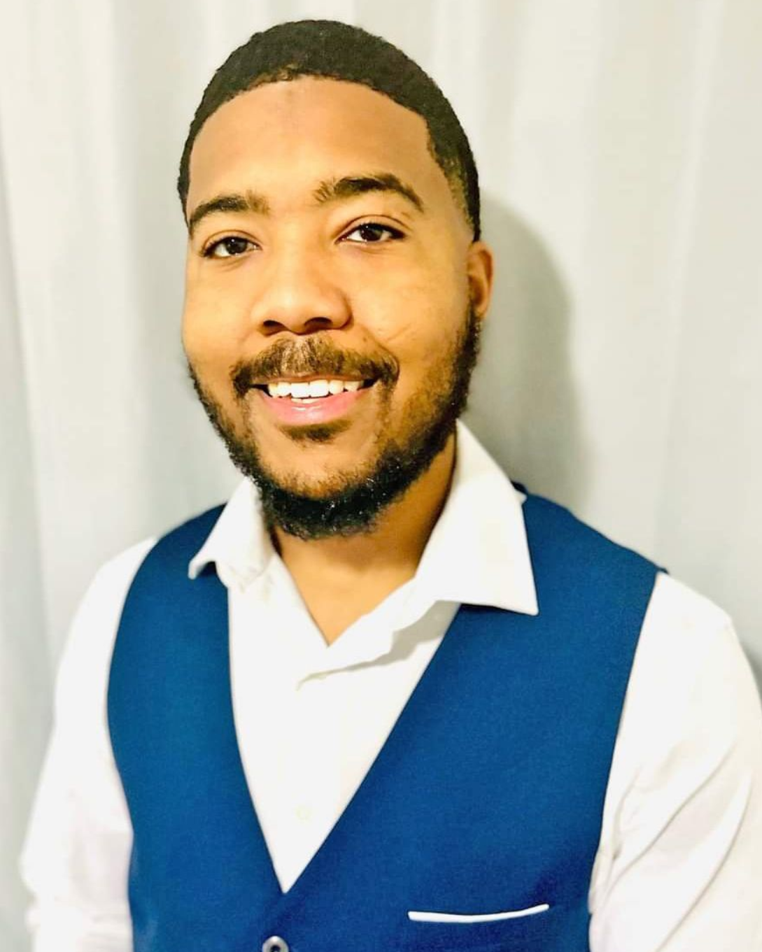 A person with a beard smiling while wearing a blue vest over a white collared shirt against a white curtain background.