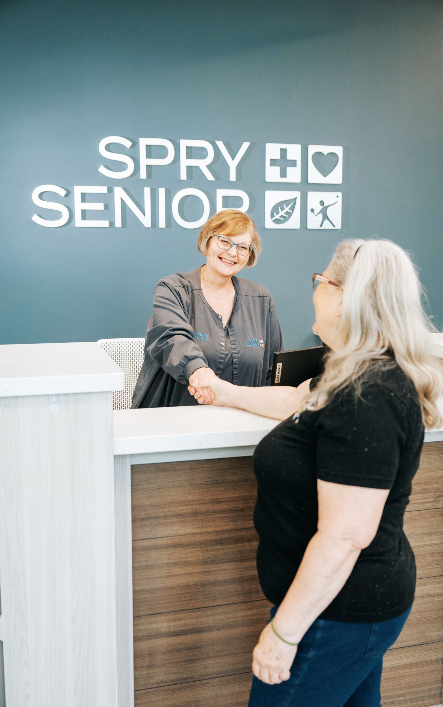 A receptionist shaking hands with a visitor at the desk of the Spry Senior center with blue walls and logo icons.