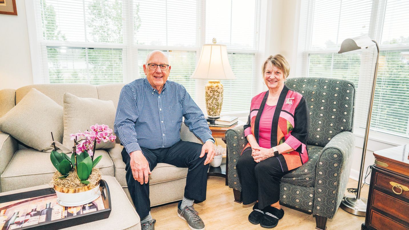 A couple sits in a bright living room, smiling at the camera in armchairs with a couch and floral arrangement nearby.