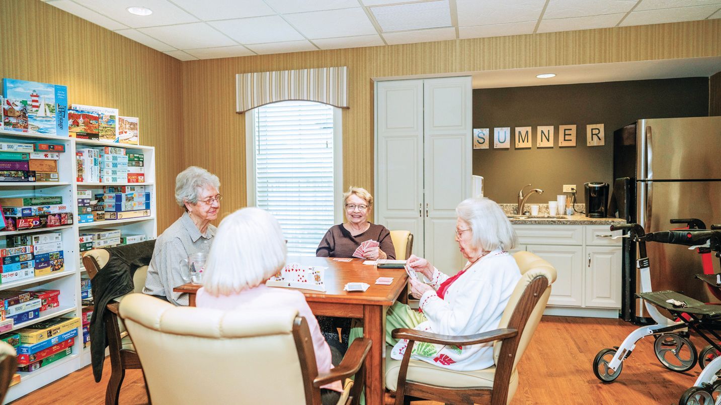 Four individuals sitting around a wooden table in a room with a game shelf, playing cards together in a community space.