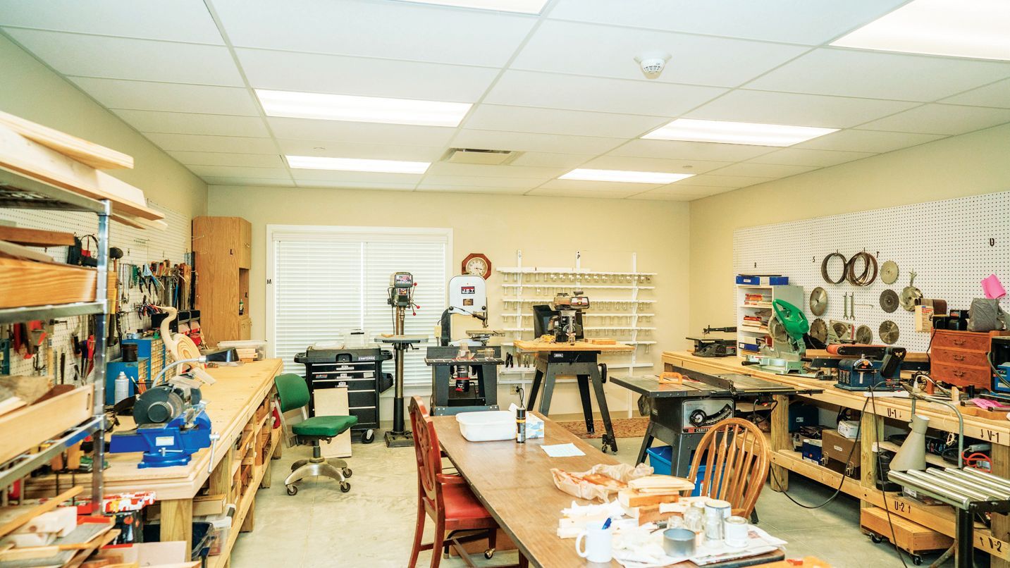 A well-organized home workshop features a central workbench, various power tools, and organized shelving on beige walls.