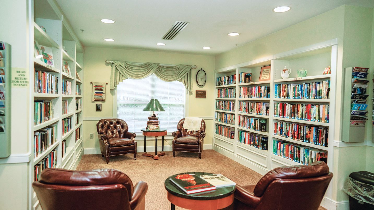 A cozy library room with two brown leather armchairs, a central round table, and bookshelves lining three walls.