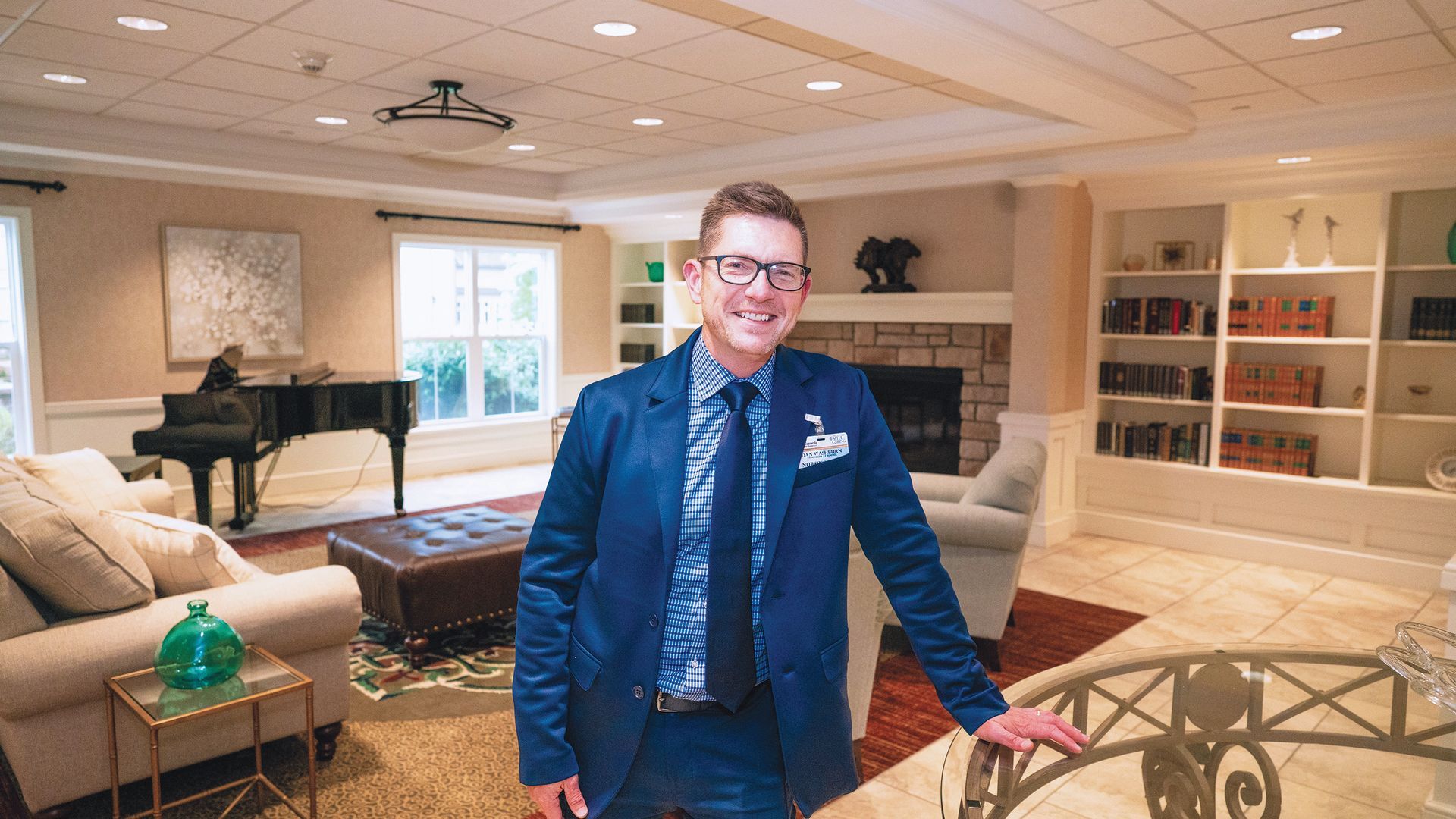 A professional smiling in a well-lit library lounge with a grand piano, fireplace, and bookshelves in the background.