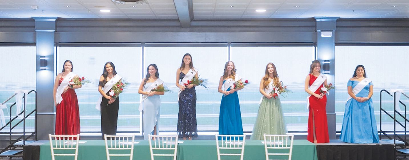 Eight contestants in formal evening gowns and sashes stand on a stage with bouquets in a brightly lit indoor venue.