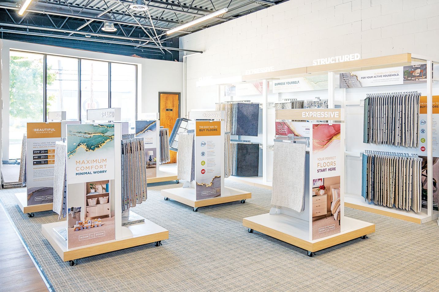 A showroom featuring multiple display stands with fabric samples and product images on a neutral-colored carpeted floor.