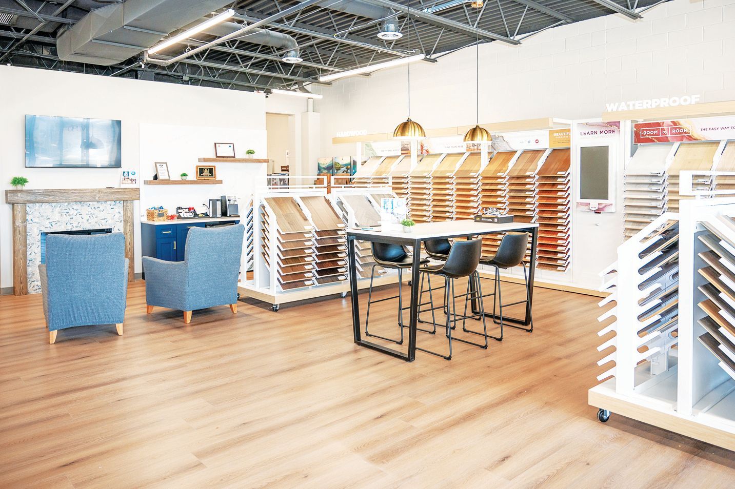 Interior showroom displaying wood flooring samples, featuring blue armchairs, a table with chairs, and wall-mounted displays.