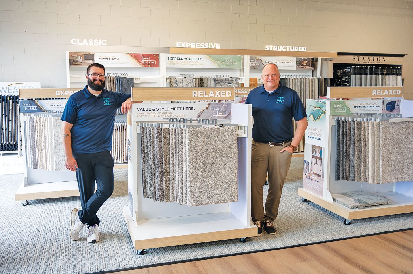 Two smiling employees in blue polo shirts stand beside carpet sample displays in a showroom.