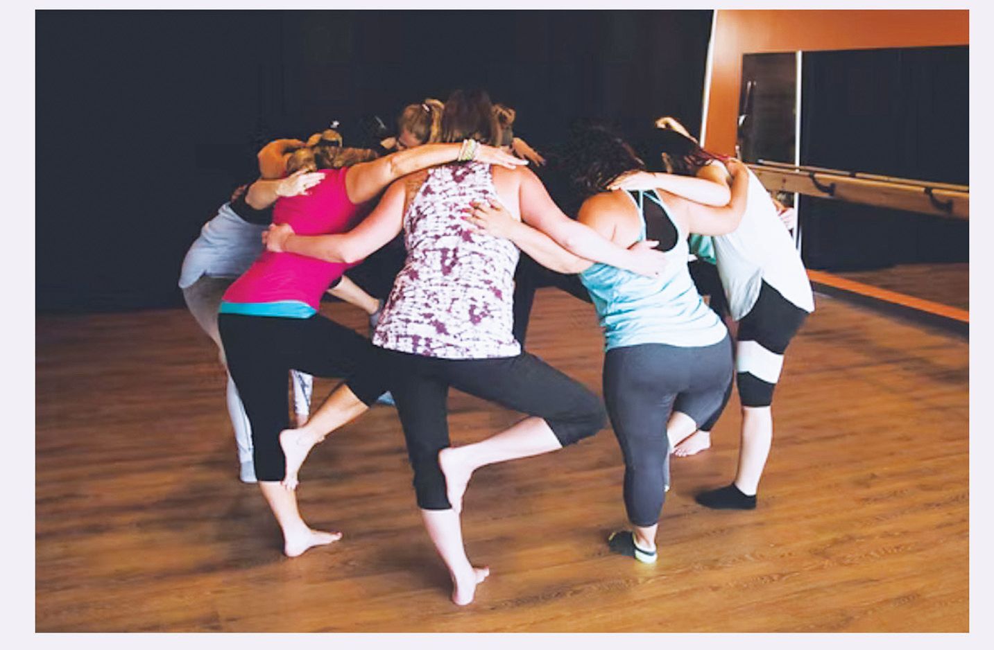 Four people in a circle balance on one leg with arms around each other's shoulders in a studio.