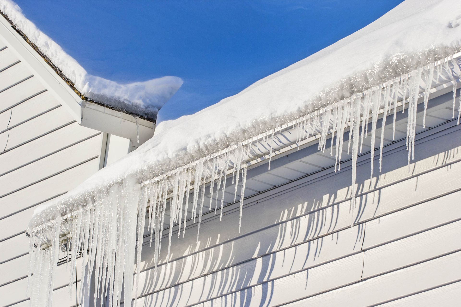 Snow-covered roof eaves with long, sharp icicles hanging from the gutter against a bright blue sky.