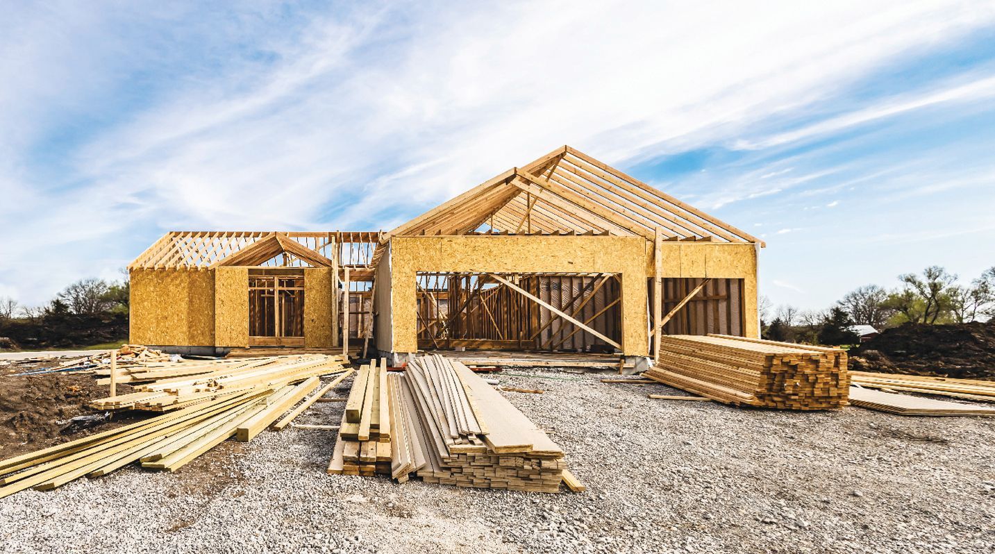 A partially constructed wooden house frame under a bright blue sky with stacks of lumber in the foreground.