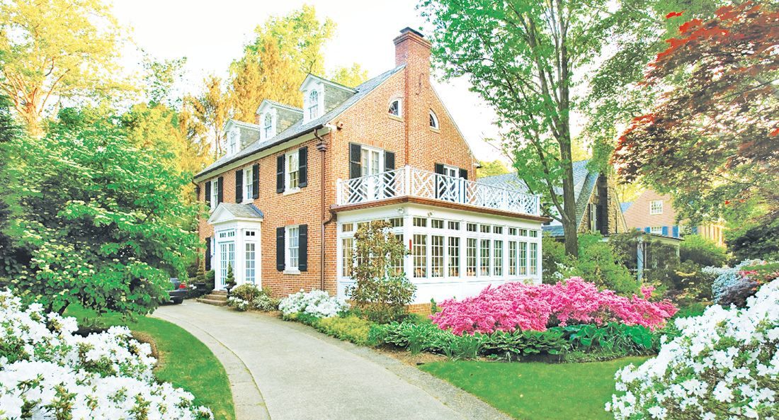 A two-story brick house with a white sunroom and dormer windows, surrounded by blooming azaleas and a paved walkway.