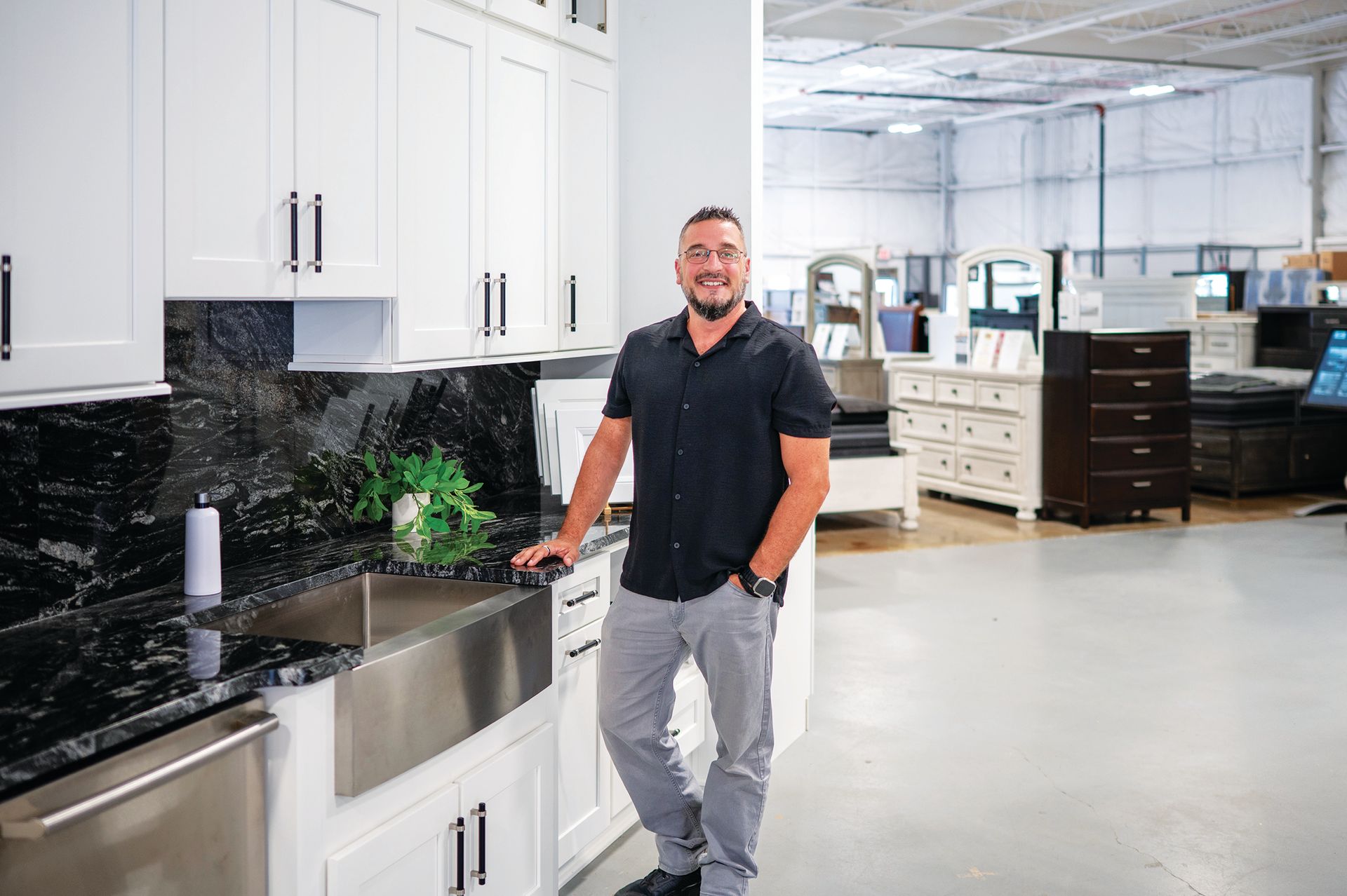 A person in a black shirt and gray pants standing next to a white kitchen cabinet display in a showroom.