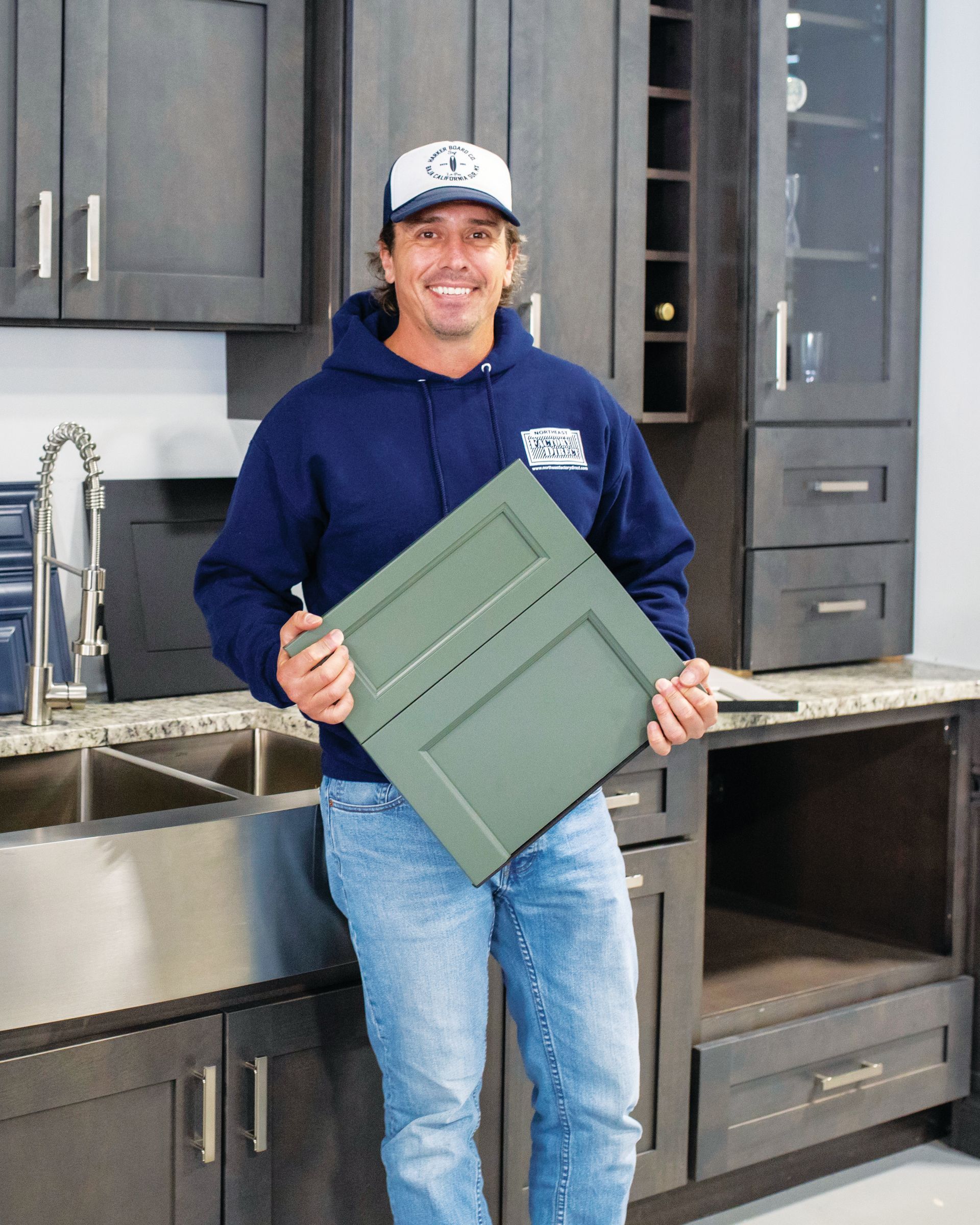 A person in a blue hoodie and cap holds a sage-green cabinet door sample in a kitchen showroom with dark cabinetry.