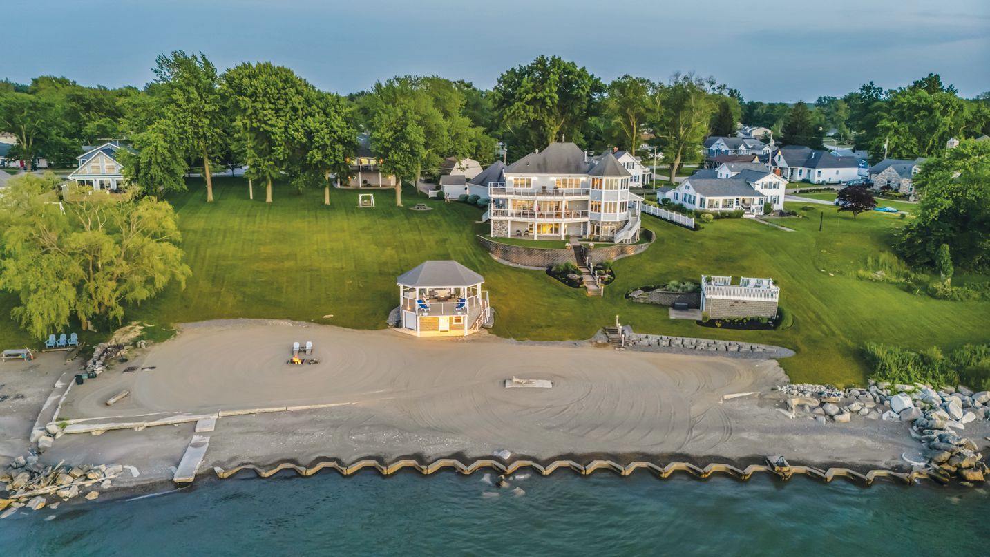 Aerial view of a lakeshore property with a beach, a gazebo, and a large house surrounded by green lawns and trees.