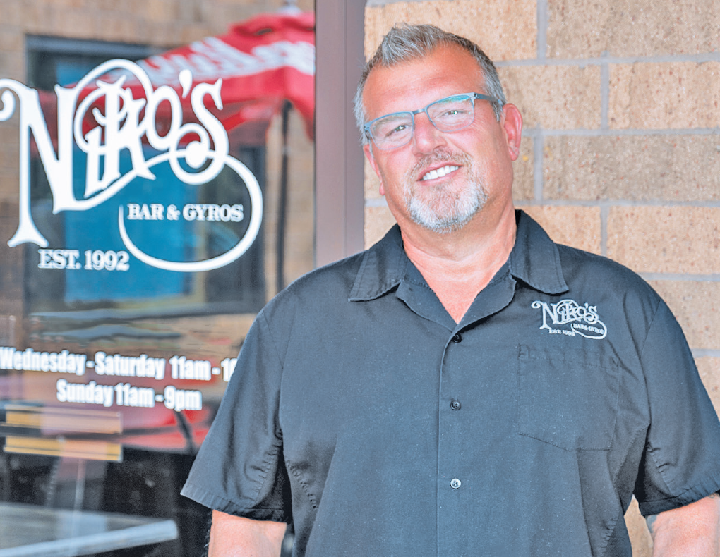 A person in a black branded shirt smiles in front of a storefront window with 