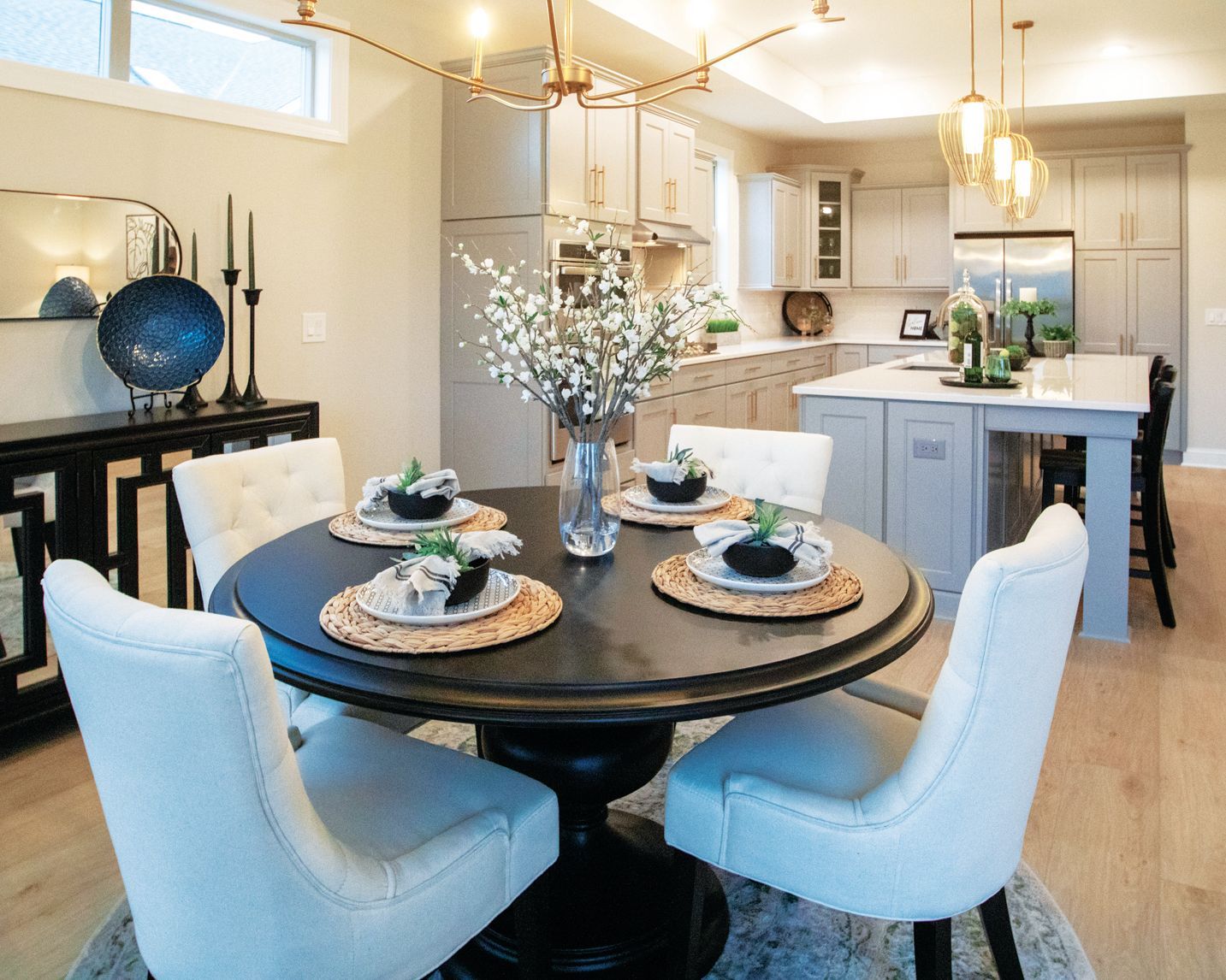 A round black dining table with white chairs in a bright, modern open-plan kitchen with light cabinets.