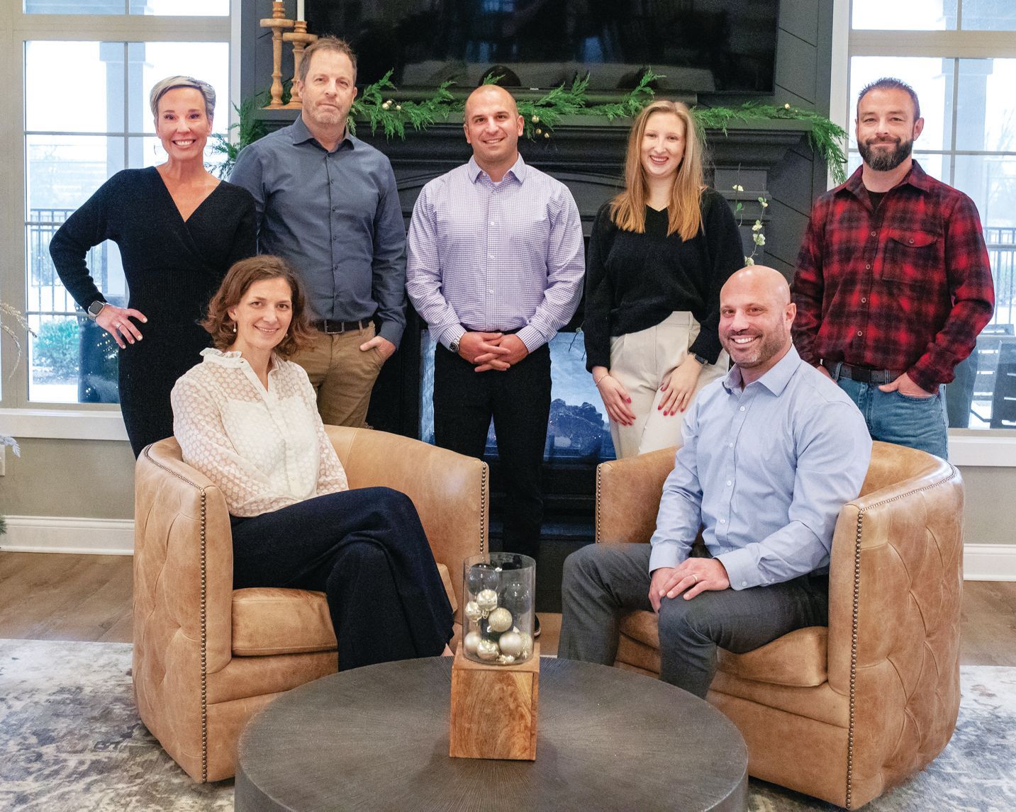 Group photo of seven people standing and sitting in a room with a fireplace and tan armchairs, smiling at the camera.