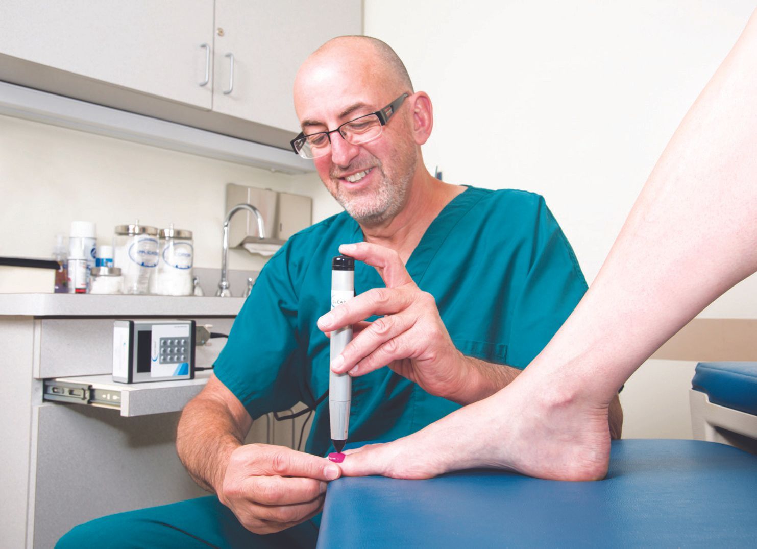 A healthcare professional in a teal scrub top uses a small medical device on a patient’s toe in a clinic office.