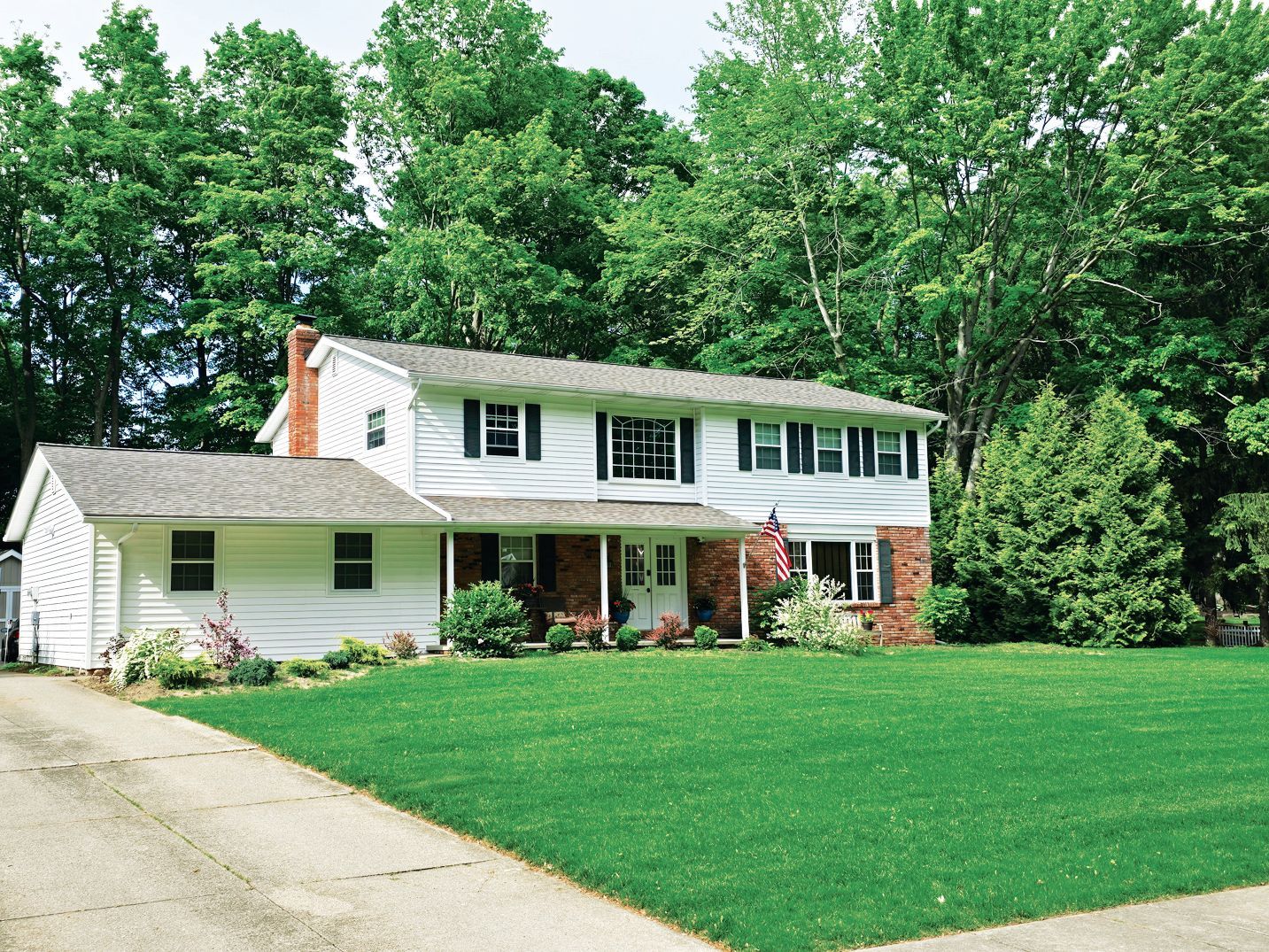 A white, two-story house with a green lawn, paved driveway, and mature trees in the background.