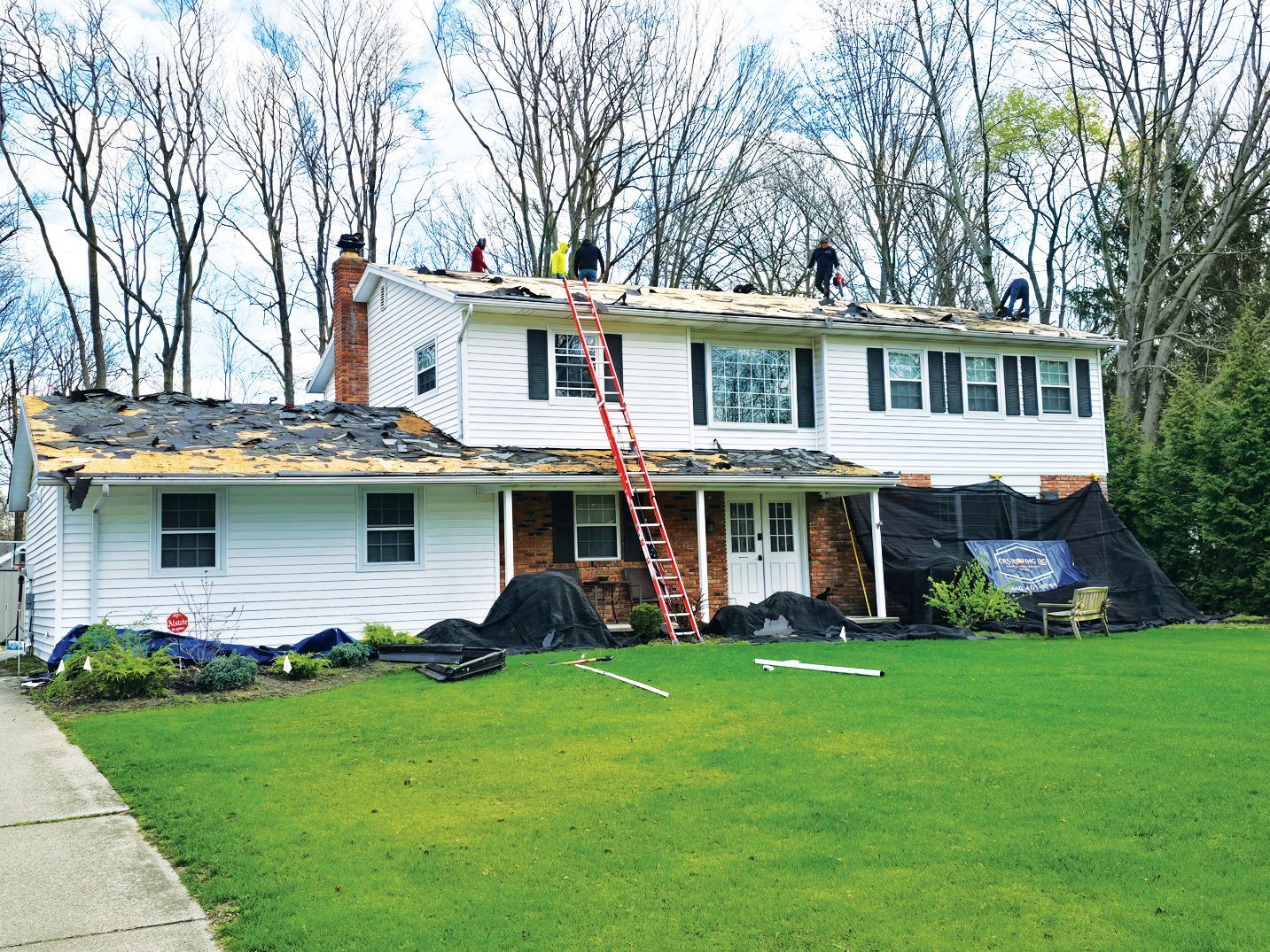 Workers repair the roof of a two-story white house with siding, using an orange ladder against the front exterior.