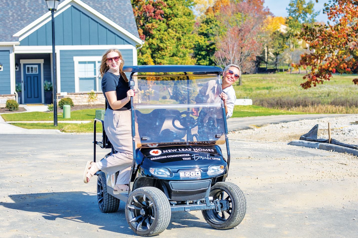 Two people pose with a black golf cart outside a blue house on a sunny day with fall foliage in the background.