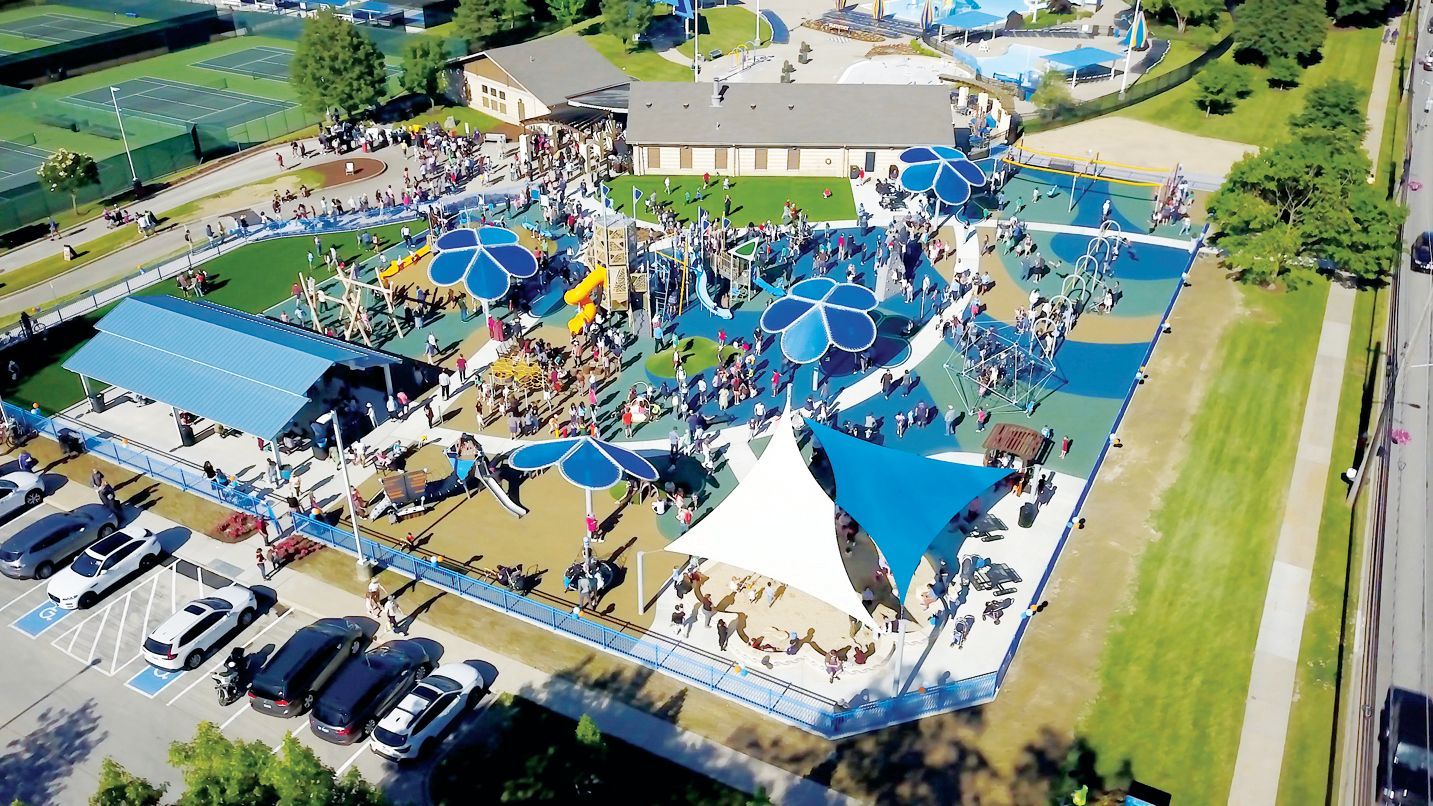 Aerial view of a crowded, colorful playground with blue shade structures, play equipment, and surrounding parking area.